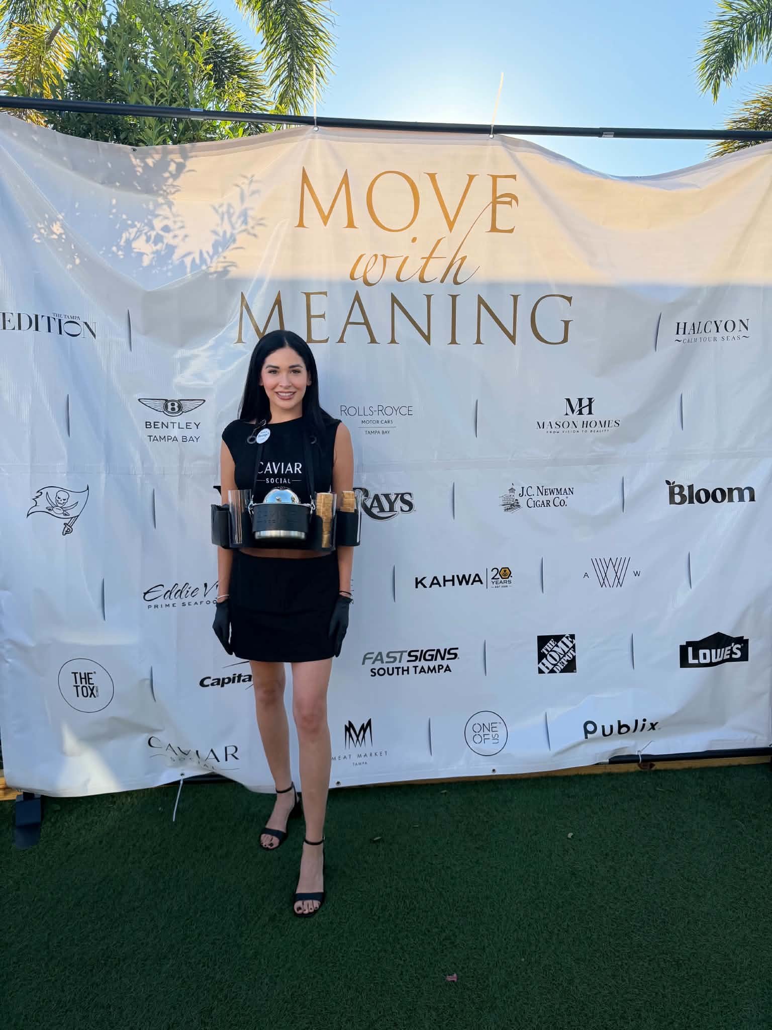 A woman in a black dress and high heels standing in front of a large banner with the words "MOVE with MEANING" and various sponsor logos. She is holding a tray with a kettle and cups, smiling at the camera outside on a sunny day.
