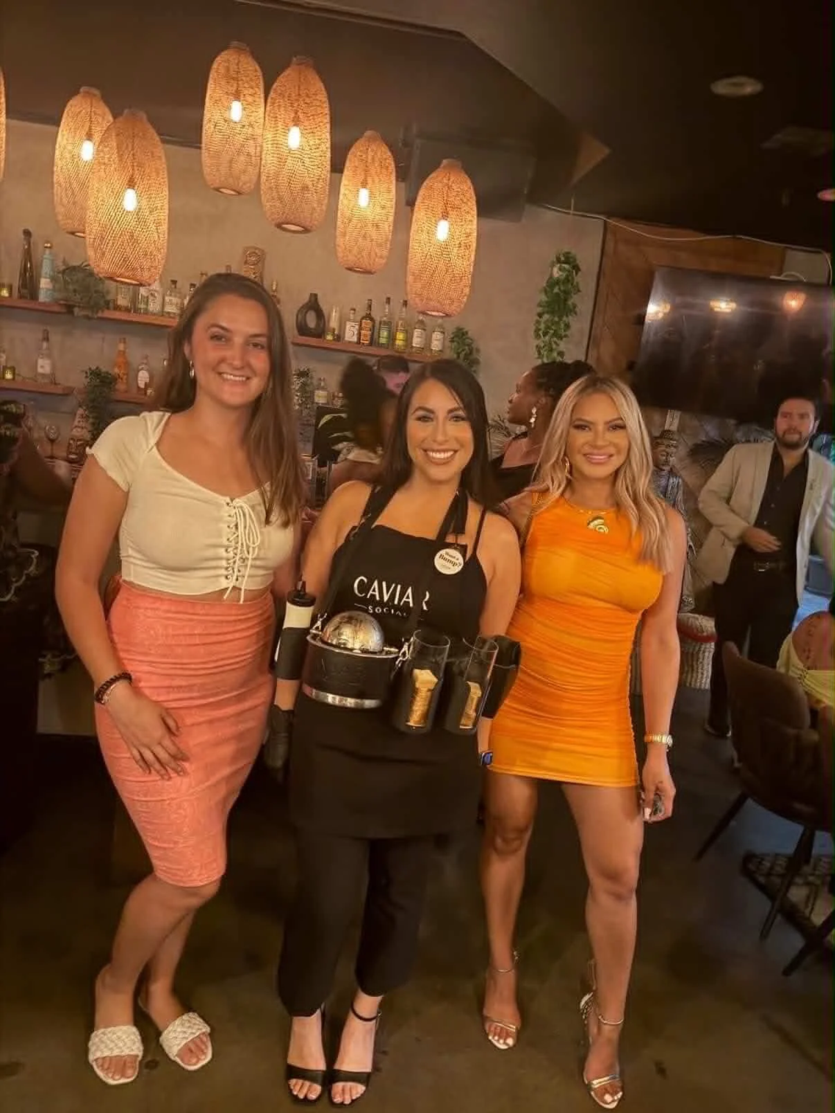 Three women standing together in a bar or restaurant, smiling and posing for a photo. The woman in the middle is a bartender wearing a black apron with bottles and bar tools attached. The woman on the left is wearing a light-colored top and pink skir