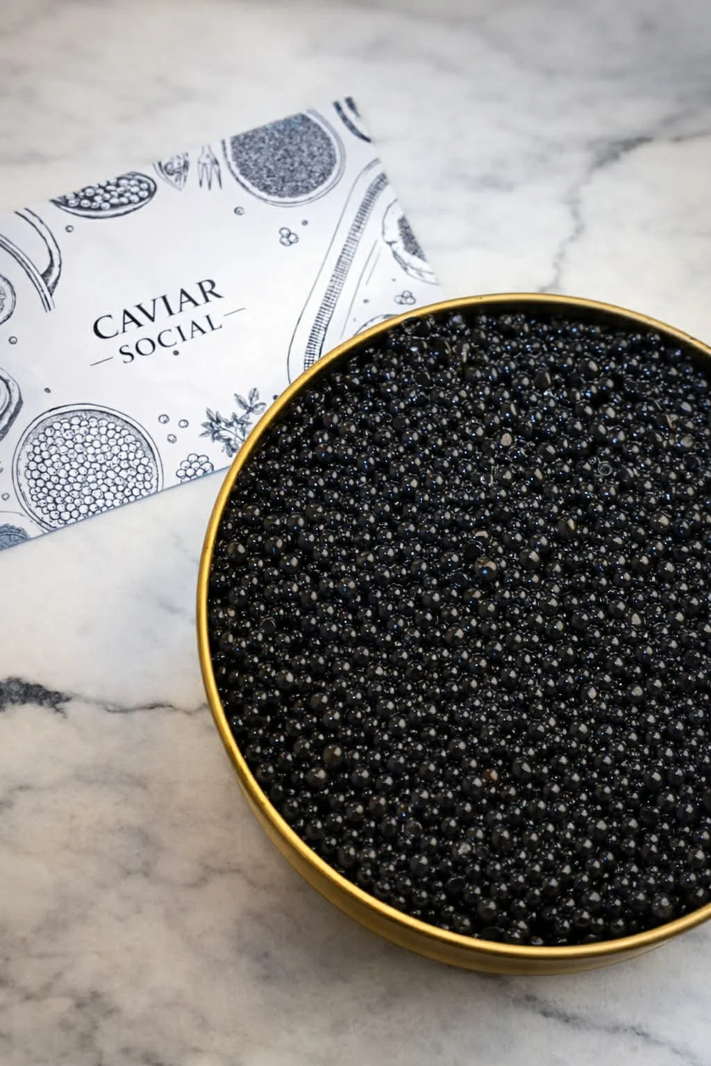 A gold-colored container filled with black caviar placed on a marble surface. A paper bag labeled 'CAVIAR SOCIAL' with decorative black and white illustrations of caviar jars is partially visible behind the container.