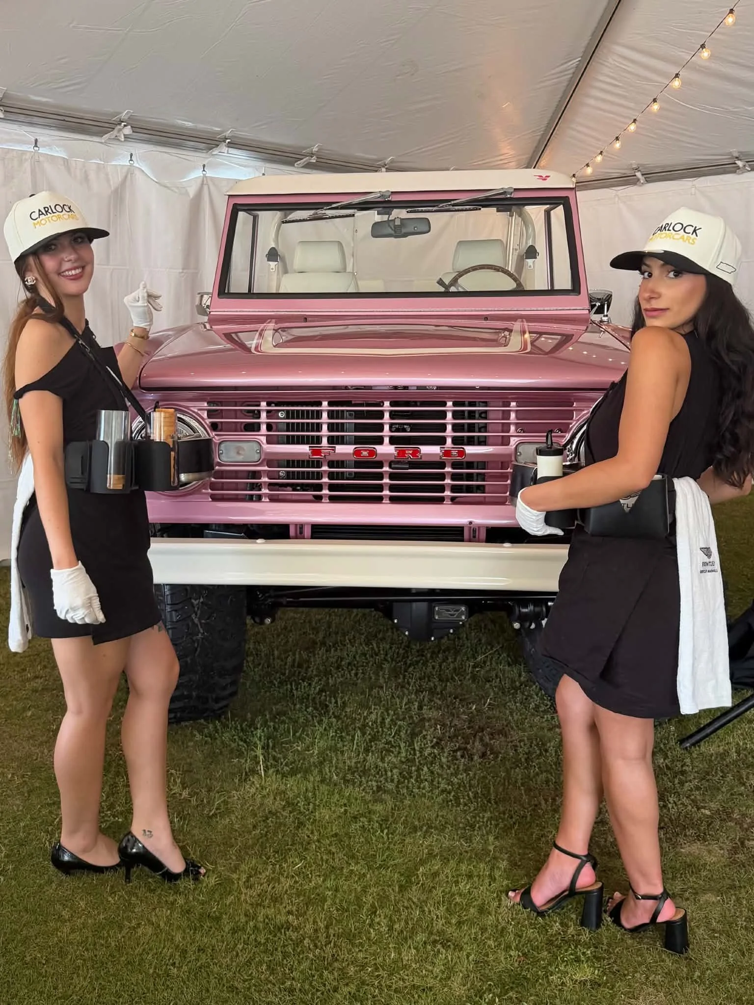 Two women in black dresses and white gloves posing in front of a pink vintage Ford vehicle under a tent with string lights.