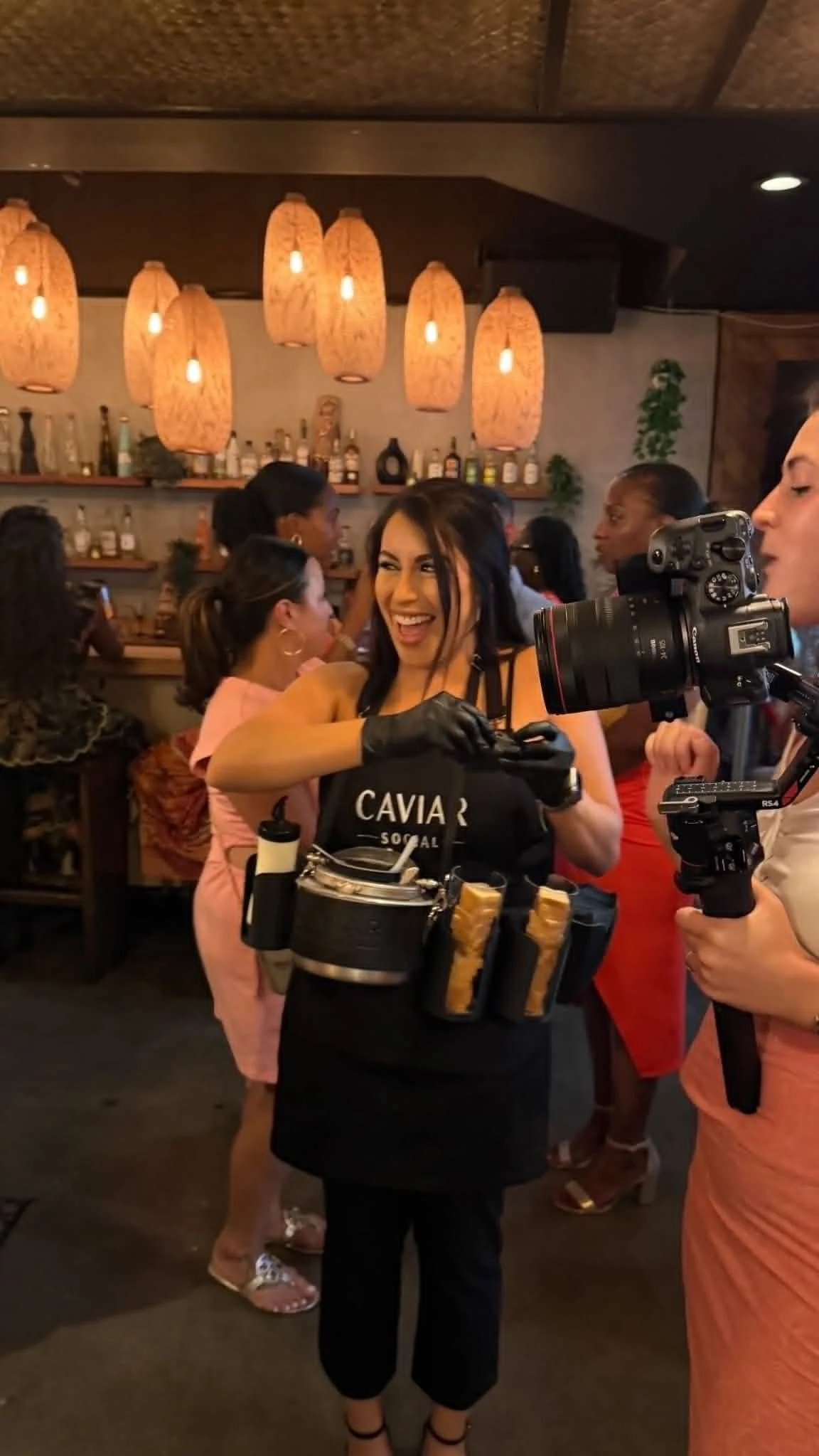 A woman working as a bartender in a restaurant or bar with a black apron that says 'Caviar Social.' She is smiling and interacting with guests, holding a black container with several breadsticks. She is also wearing black gloves and a black apron wit