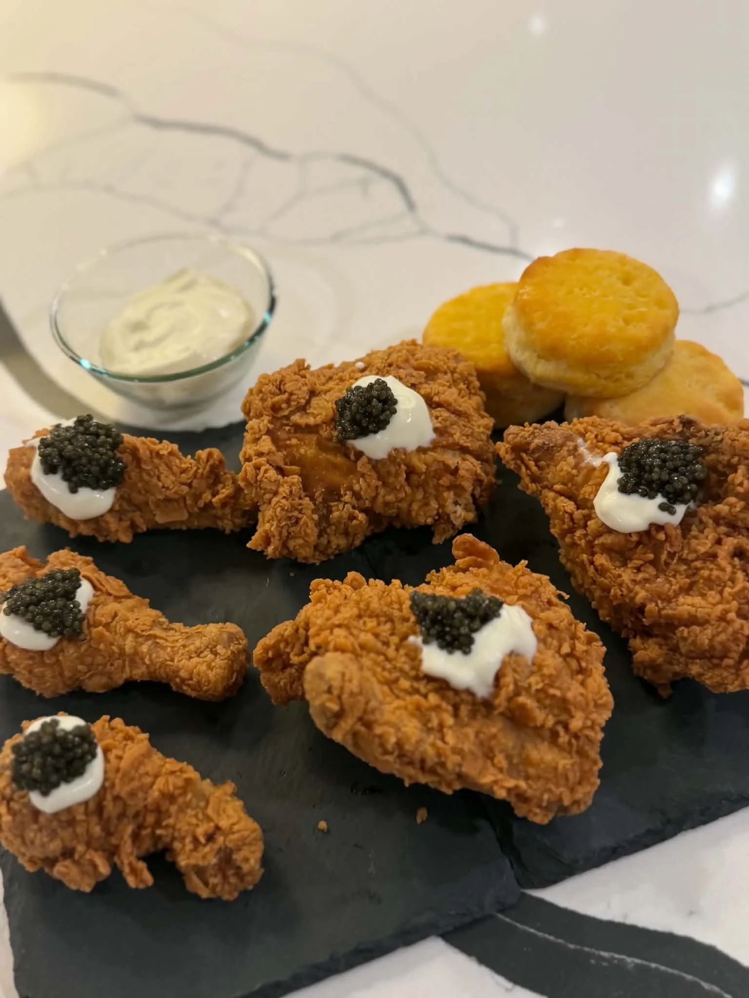Crispy fried chicken drumsticks with dollops of mayonnaise topped with black caviar, served on a black slate platter with a side of fried potato rounds and a small bowl of creamy sauce.