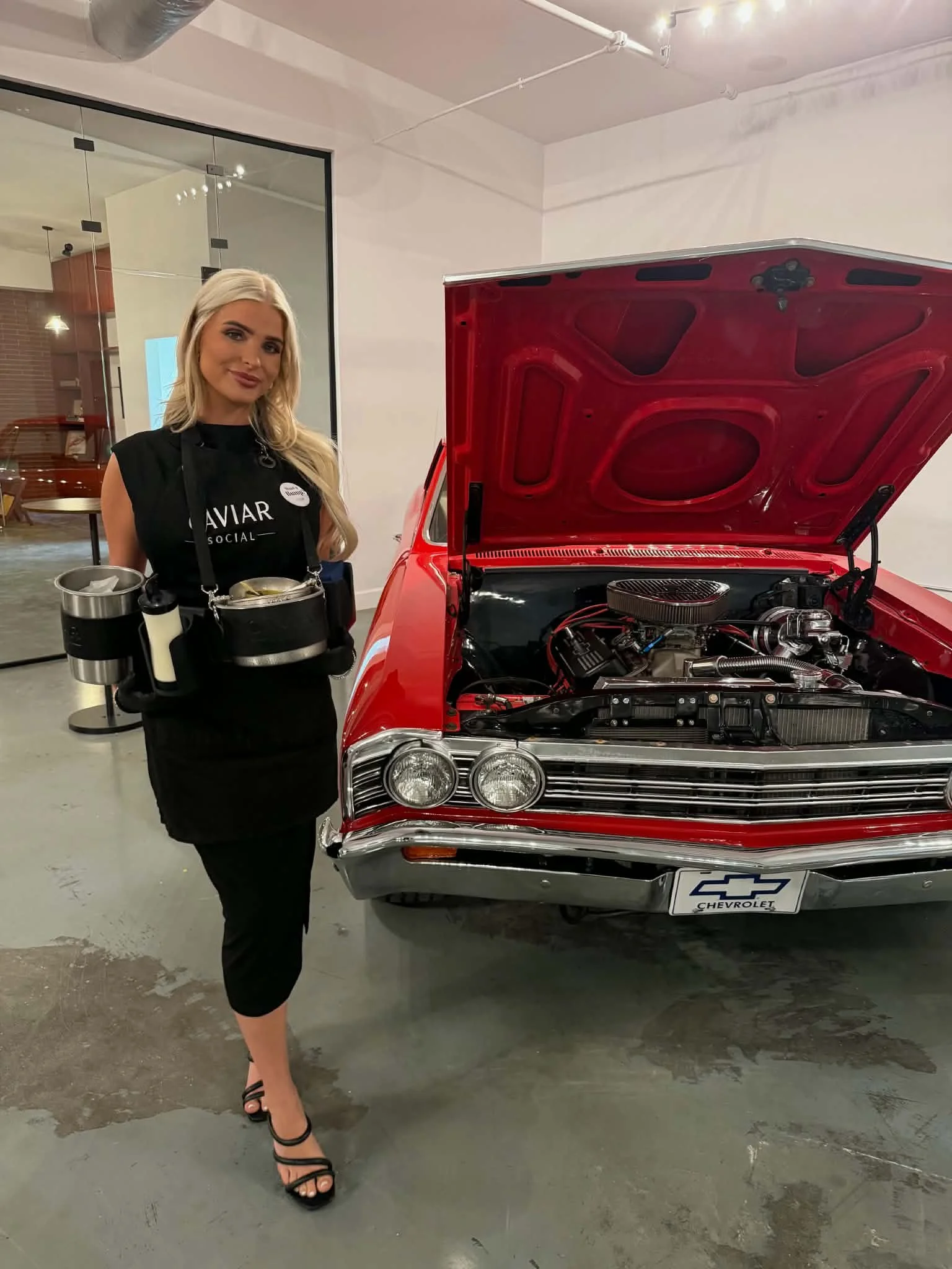 A woman dressed in black standing next to a red classic Chevrolet car with its hood open, showing the engine. She is wearing a black dress and high heels, and holding a tray with coffee cups.