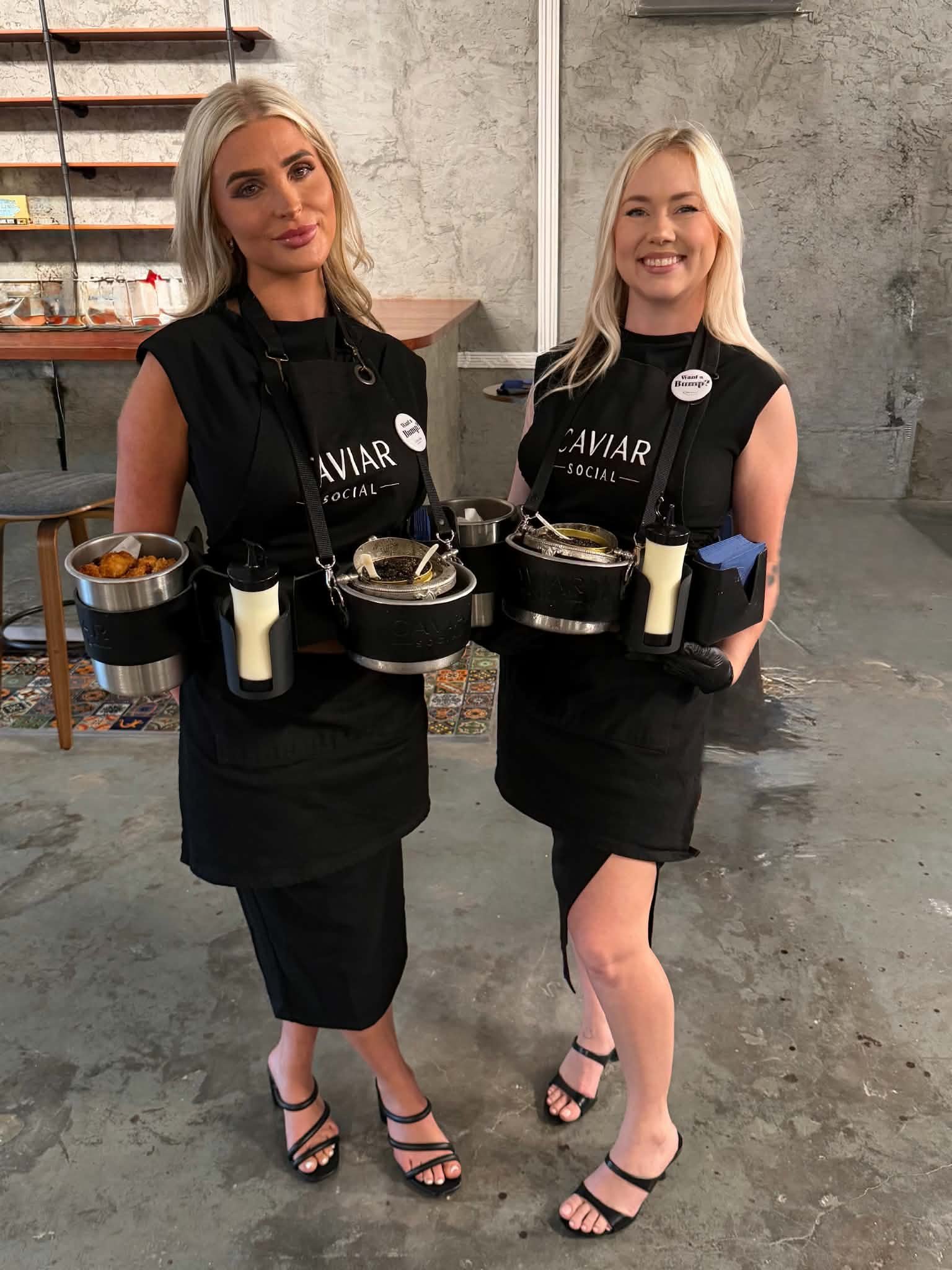 Two women dressed in black dresses and high heels, holding trays with caviar, standing on a concrete floor in an indoor setting with gray walls.