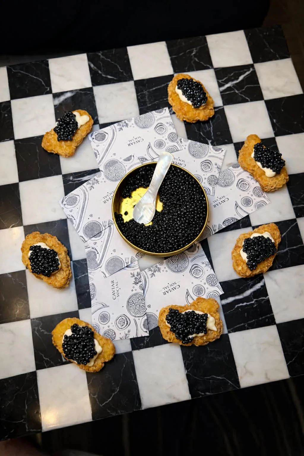 A black and white checkered marble table with five pieces of fried food topped with black caviar and dollops of white sauce, a bowl filled with black caviar and a small silver spoon.