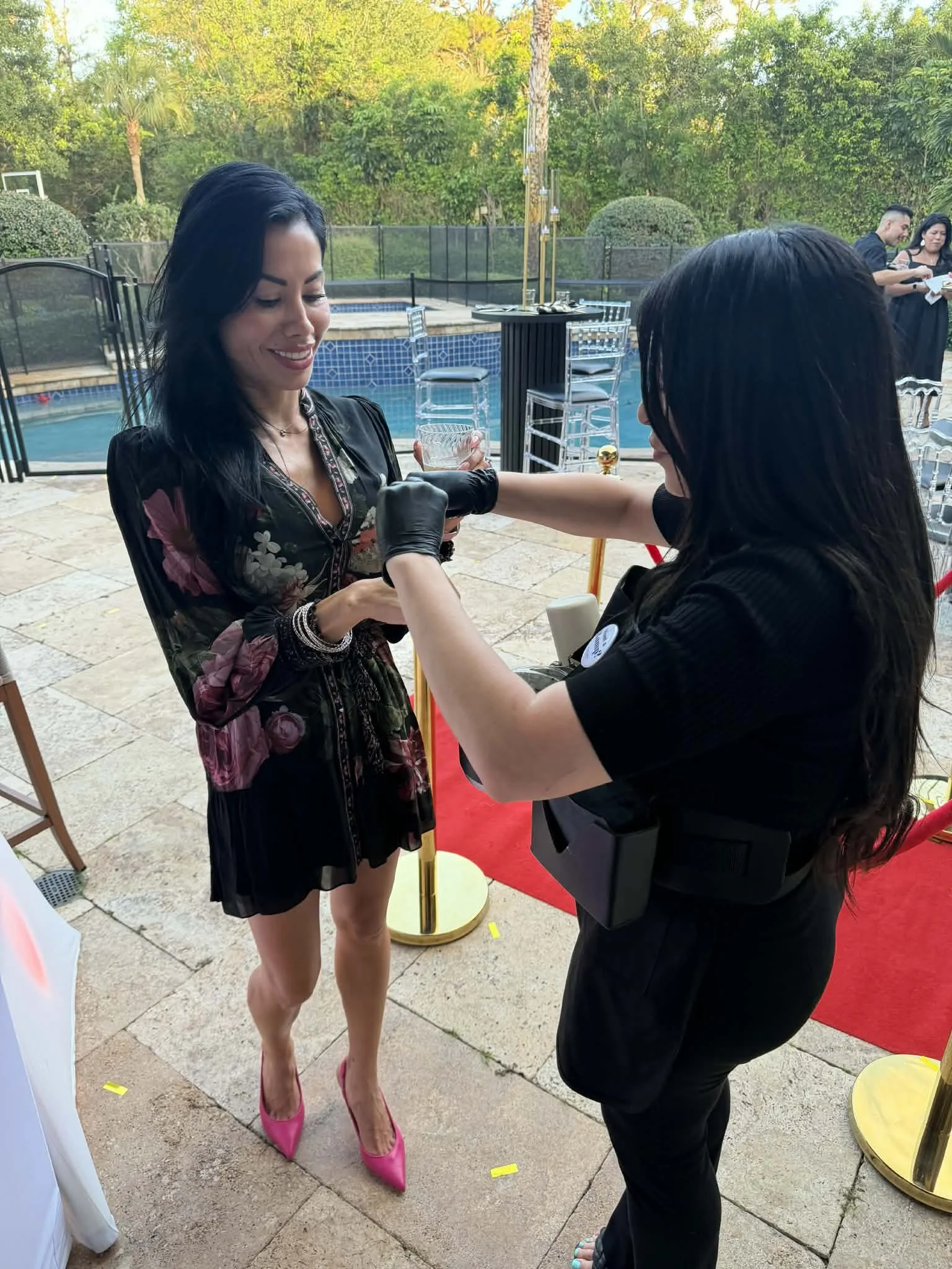 A woman with black hair and pink high heels receives a vaccination from a healthcare worker at an outdoor event by a pool.