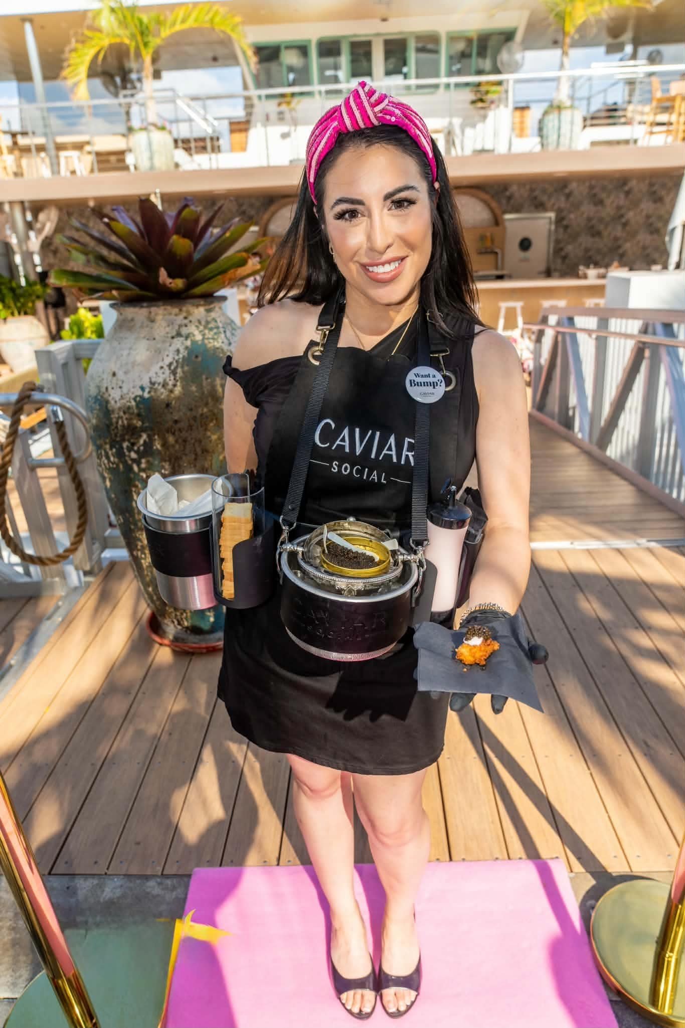 A smiling woman with dark hair wearing a pink striped head wrap, black off-shoulder top and apron holding a tray with caviar and a small dish of orange flowers. She is standing on a pink yoga mat on a wooden deck with outdoor furniture and potted plants in the background.