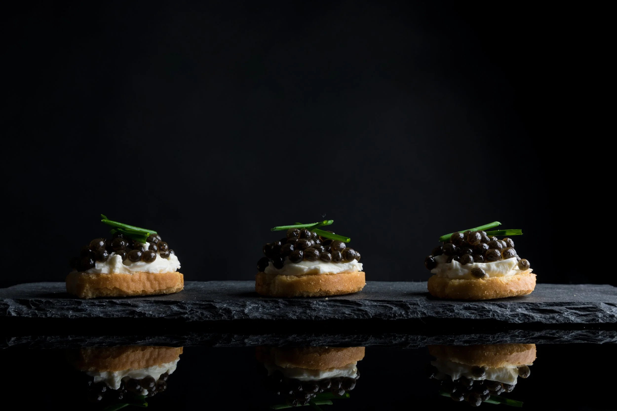 Three canapés with a biscuit base, topped with creamy white spread, black caviar, and a small green garnish, arranged on a black slate surface against a dark background.