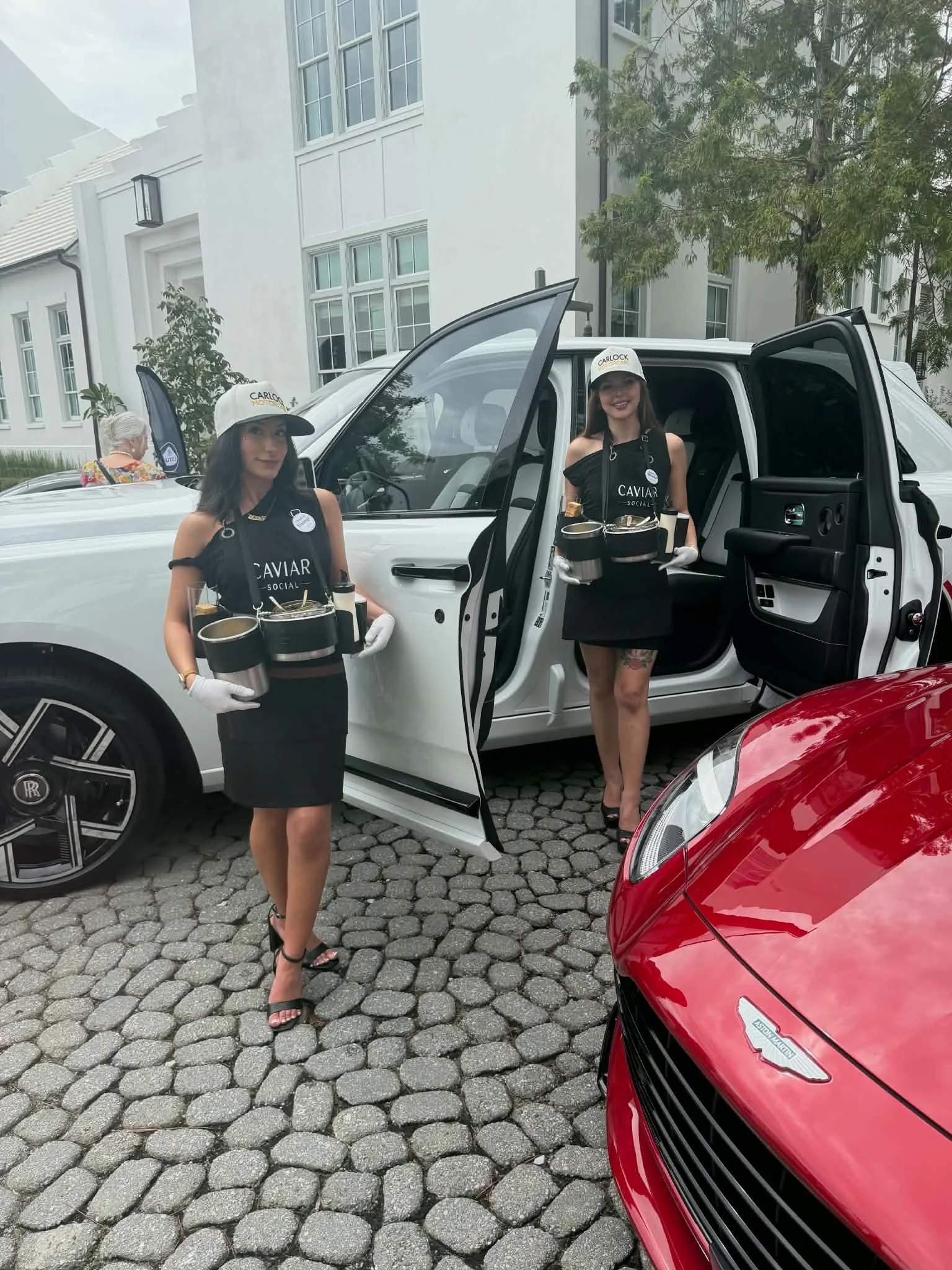 Two women dressed in black with white gloves, hats, and aprons reading 'Caviar Social' are standing beside an open white electric vehicle, holding trays with caviar jars. One woman is leaning against the car, and the other is standing near the open d