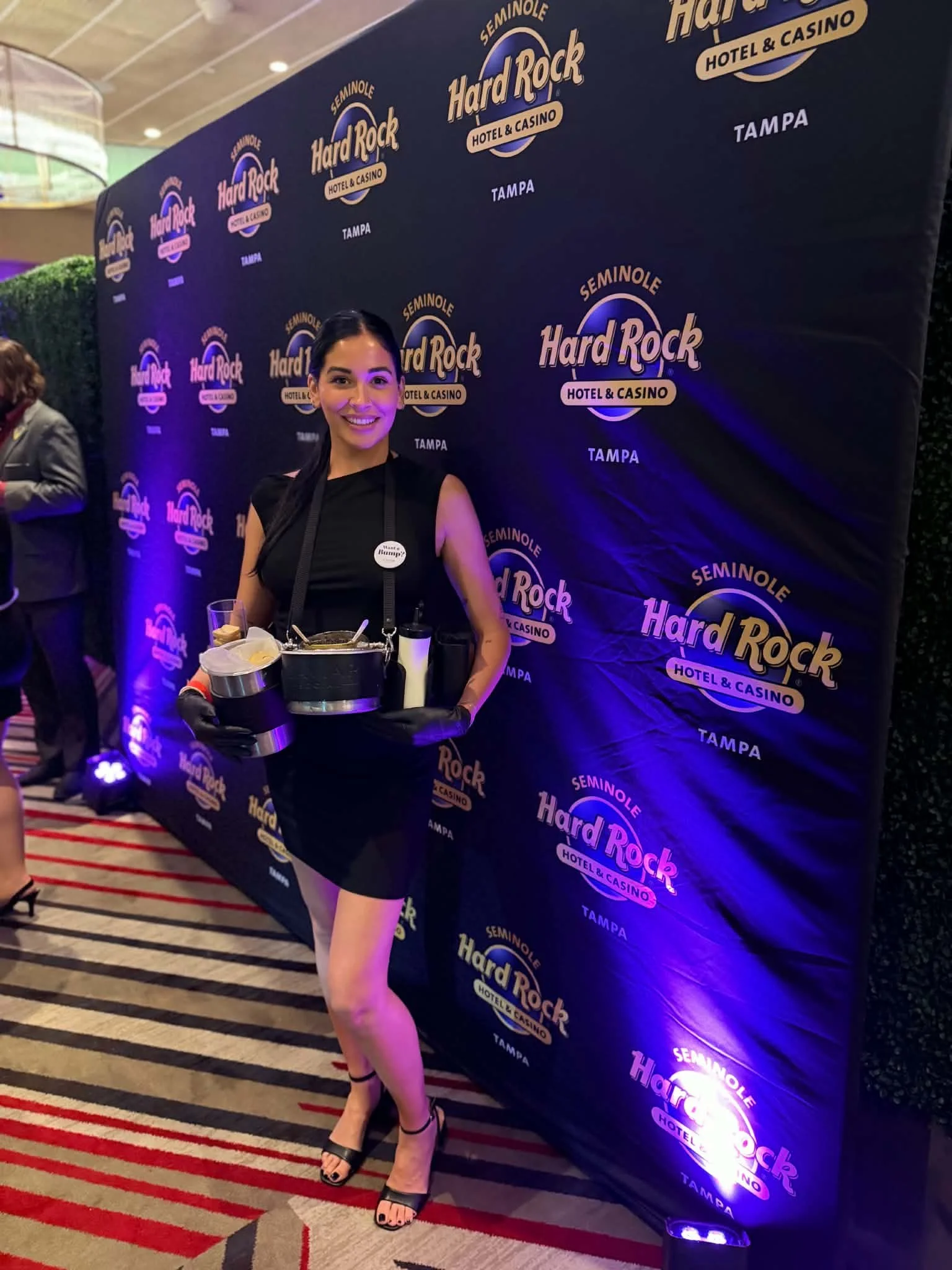 A woman standing in front of a step-and-repeat banner with Hard Rock Hotel & Casino and Tampa logos, holding a tray with food. She is dressed in a black dress and high heels, smiling for the camera at an event.