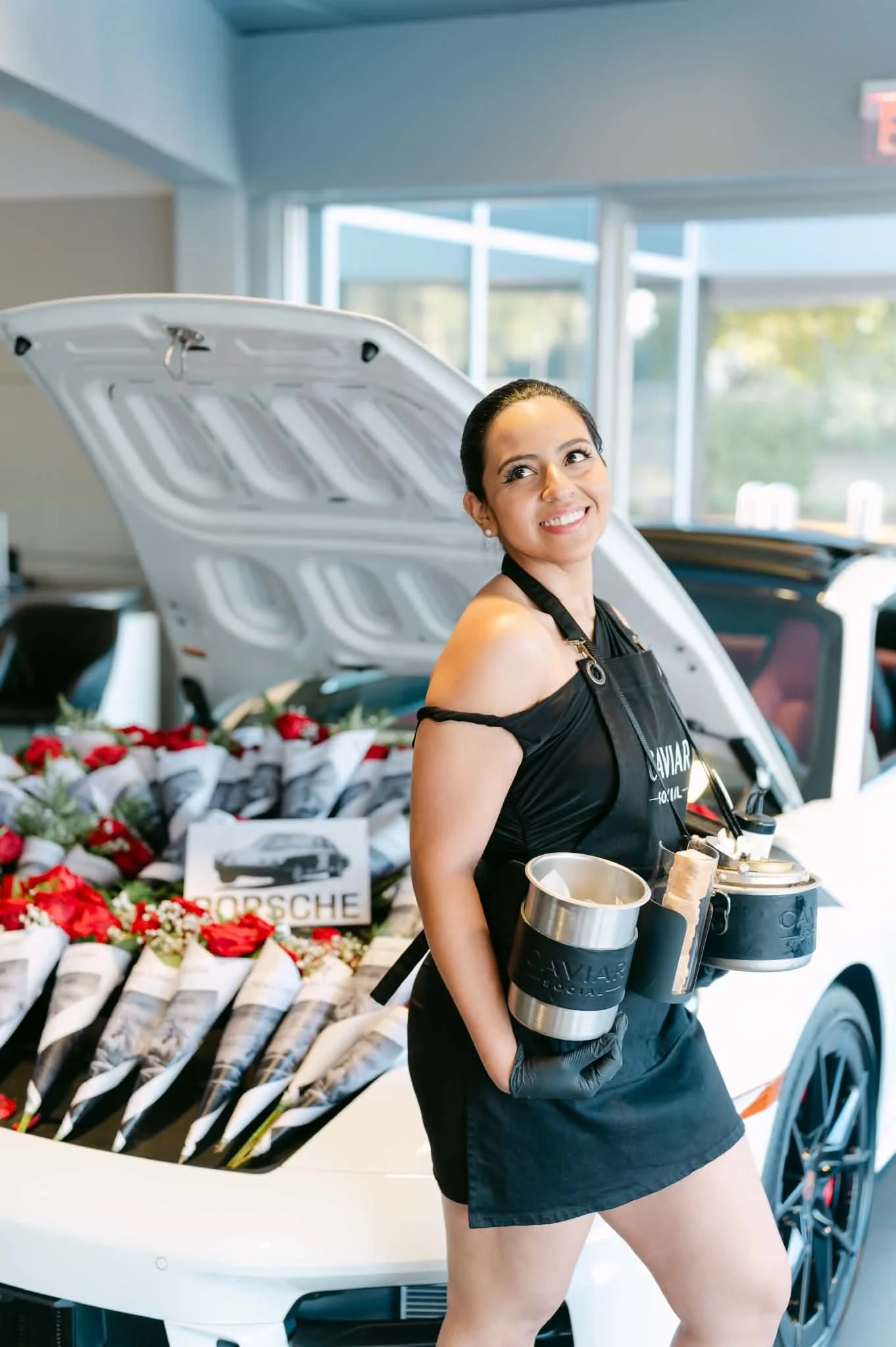 A smiling woman wearing an apron and black gloves standing in front of an open car hood with a Porsche logo, surrounded by flower bouquets in white cones, inside a modern building with large windows.