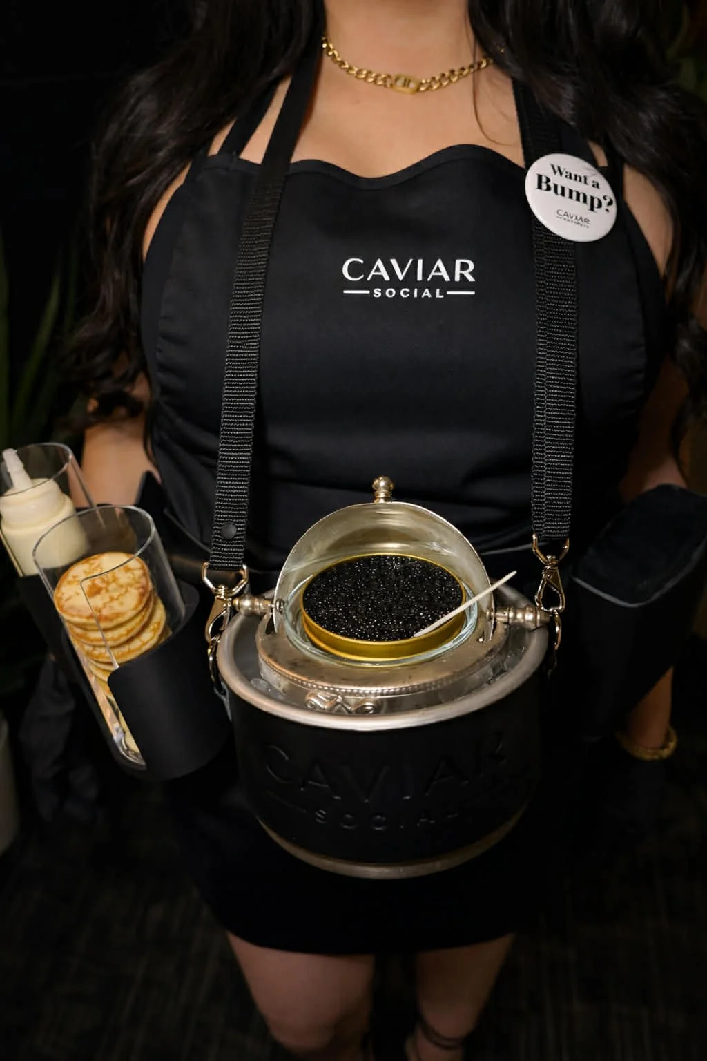 A woman wearing a black apron with 'CAVIAR SOCIAL' printed on it, holding a container with black caviar, and a small spoon. She is also wearing a gold chain necklace and has a button on her apron that says 'Want a Bump?'.
