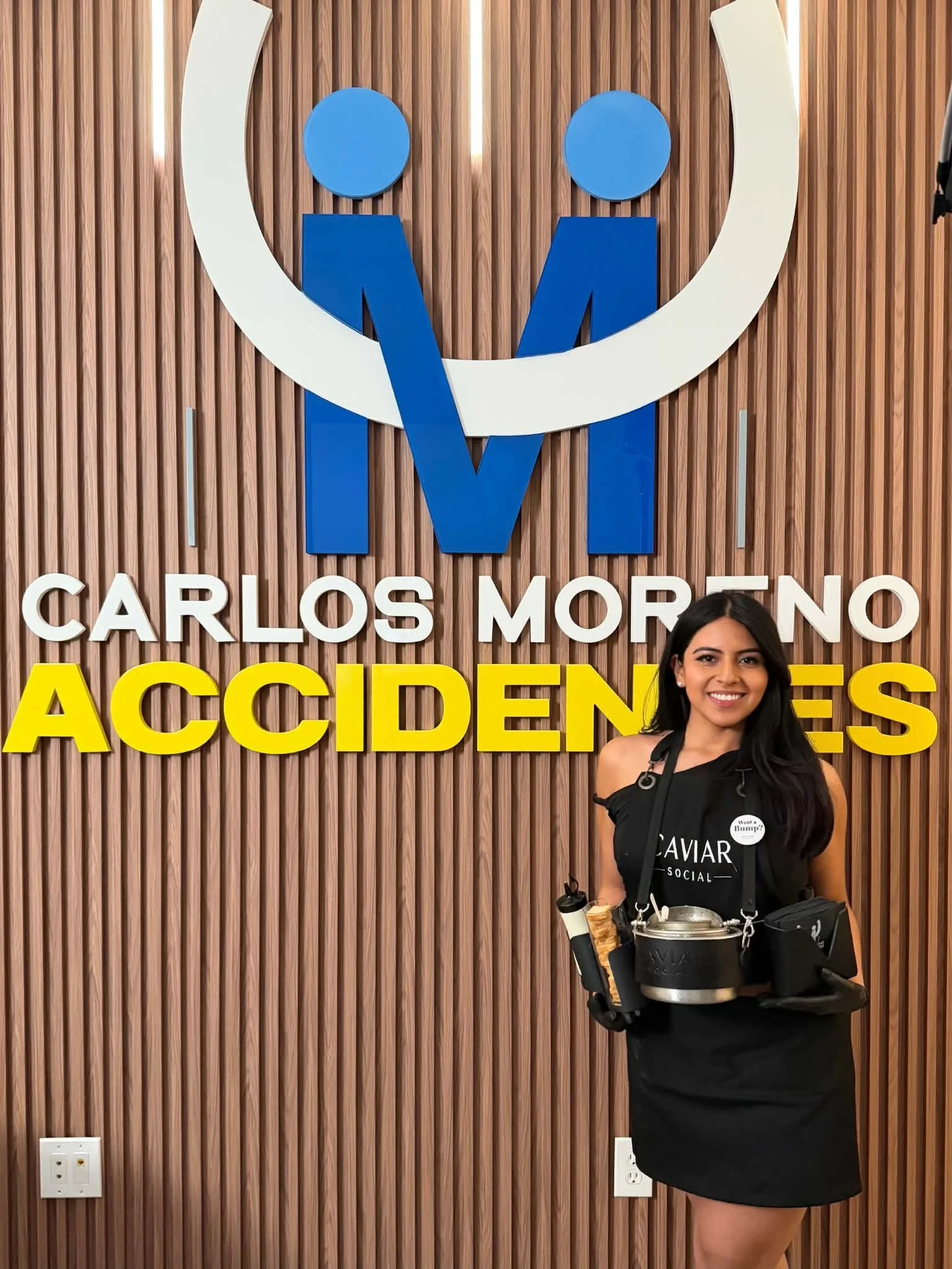 A woman is standing in front of a wooden wall with a logo and text for 'Carlos Moreno Accidents.' She is holding a tray with food and drinks and wearing a black dress with a 'Caviar Social' apron, a button, and a wallet or small bag hanging from her 
