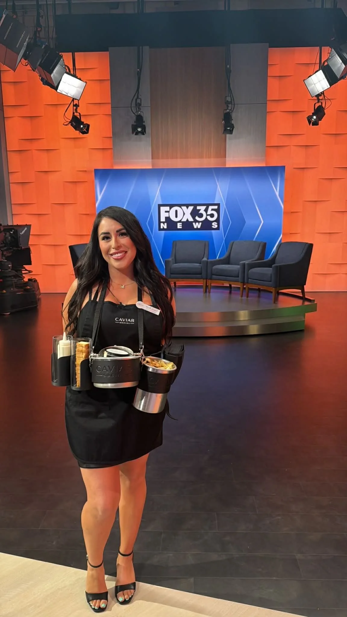 A woman standing in a TV studio with a FOX 35 NEWS backdrop and three chairs behind her, holding a tray with snacks and beverages.