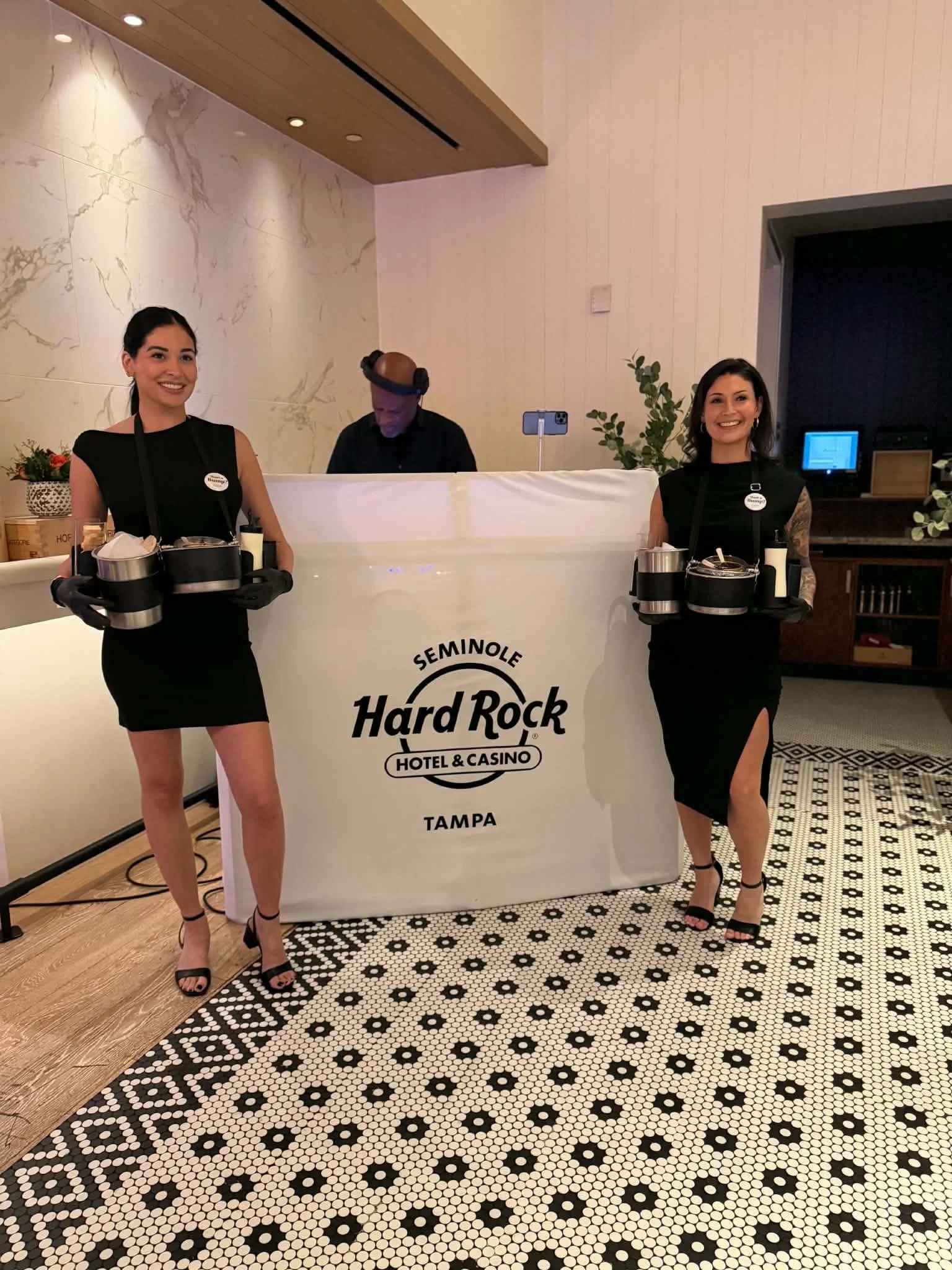 Two women in black dresses and high heels standing on patterned black and white floor, holding trays with utensils and condiments, at a reception desk with "Hard Rock Hotel & Casino Tampa" logo. Two people are behind the desk, one is focused on their