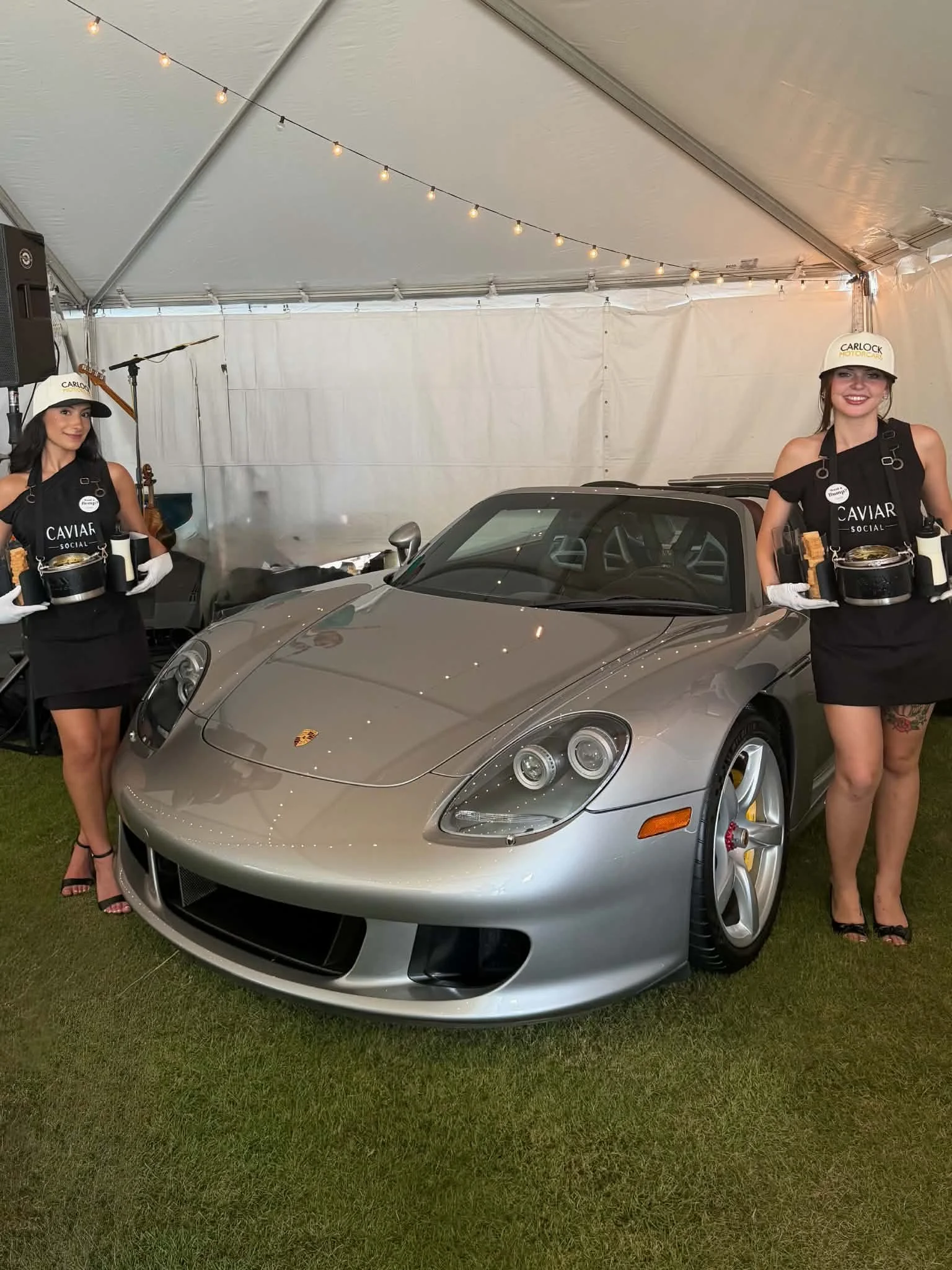 Two women dressed in black dresses, wearing white hard hats with 'CARLOCK' written on them, are standing next to a silver sports car inside a large white tent. They are smiling and holding trays with food and drinks.