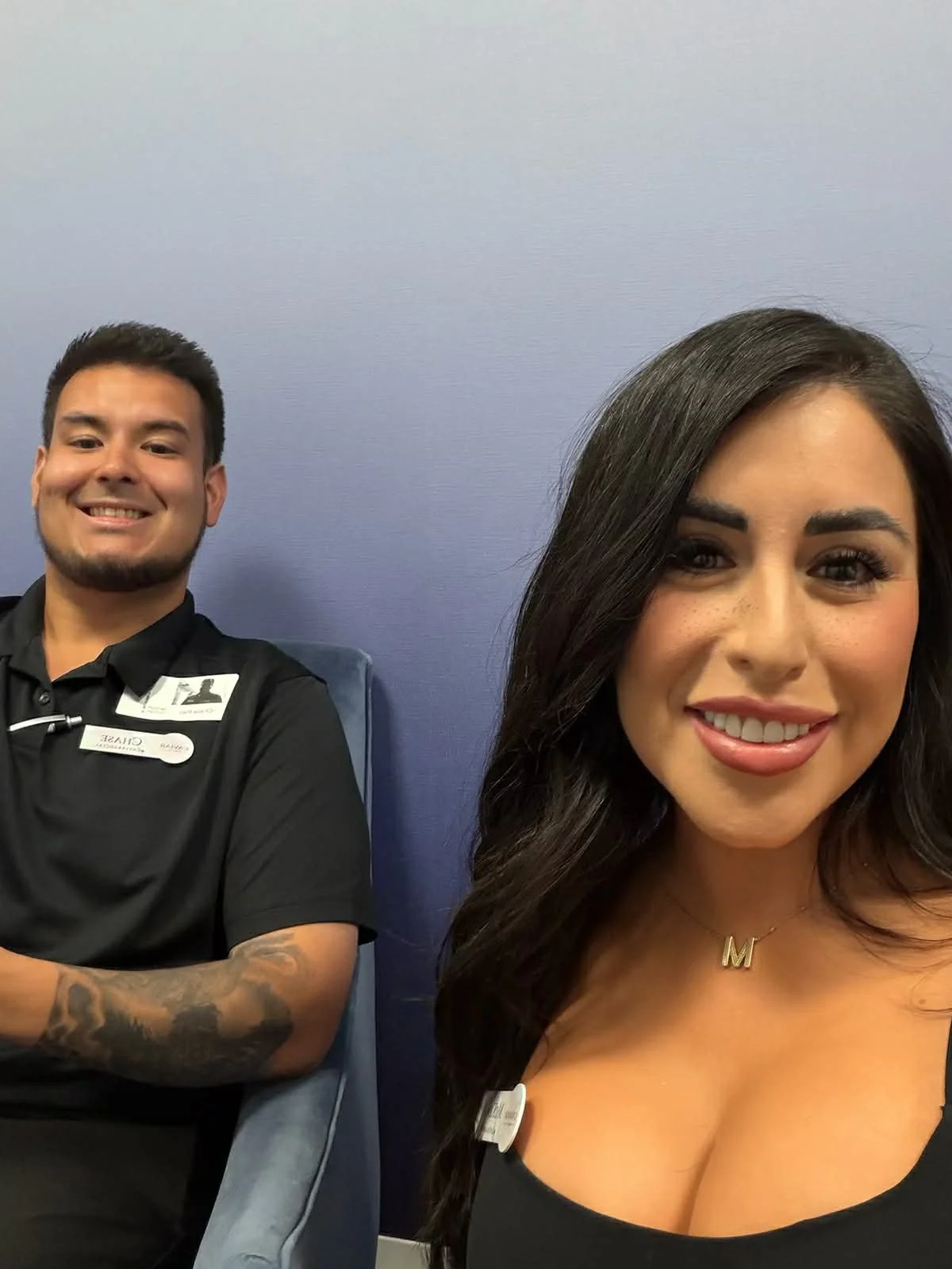 A woman taking a selfie with a man sitting behind her in a blue chair, both smiling. The woman has dark hair, wearing a black top with a name tag, and a gold necklace with the letter 'M'. The man has short dark hair, facial hair, and is wearing a bla