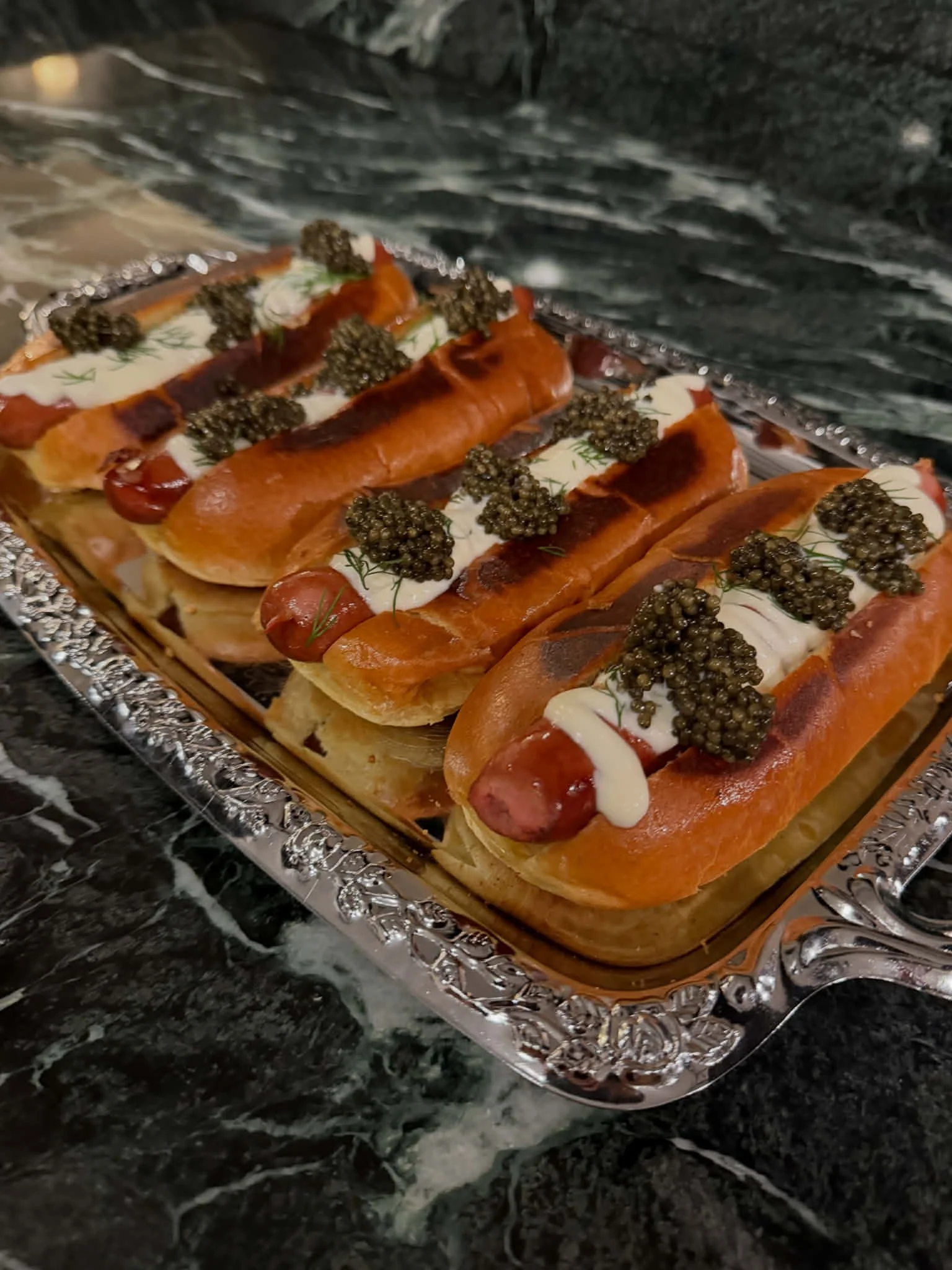 Open-faced hot dogs on a silver tray topped with sour cream, caviar, and herbs, sitting on a dark marble surface.