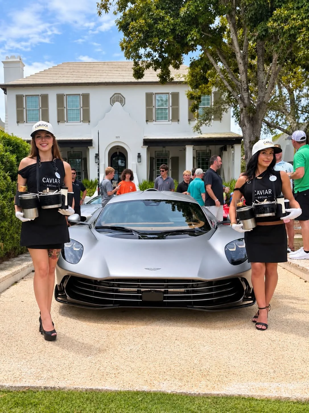 Two women dressed as servers holding trays with cans and cups stand on either side of a silver luxury sports car parked in front of a large white house with a crowd of people in the background.