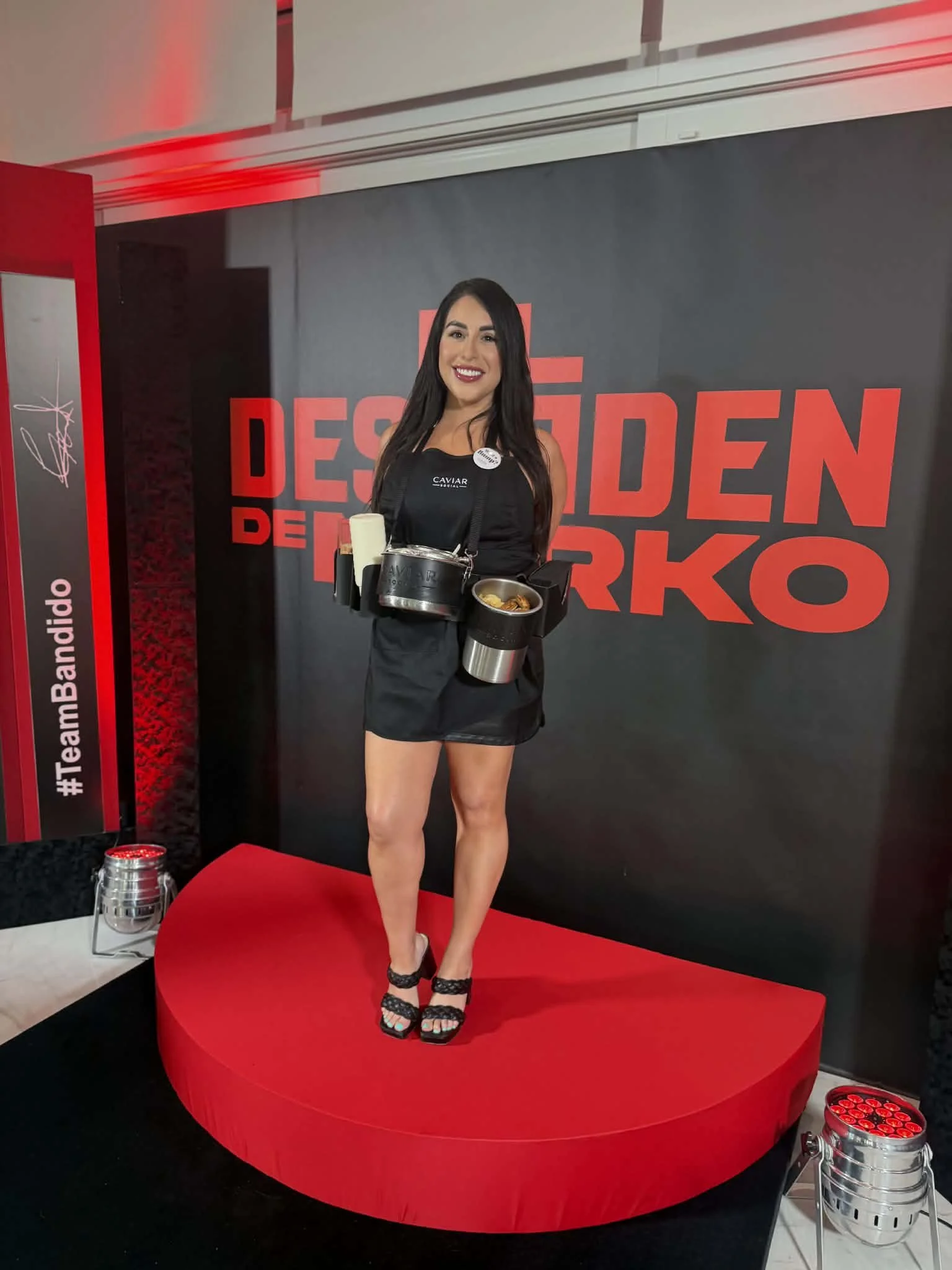 A woman with long black hair in a black dress and heels standing on a red circular stage at an event with a black and red backdrop that has the words 'Design' and 'Darko' visible and hashtag '#TeamBanda' on a vertical banner to the side. She is smili