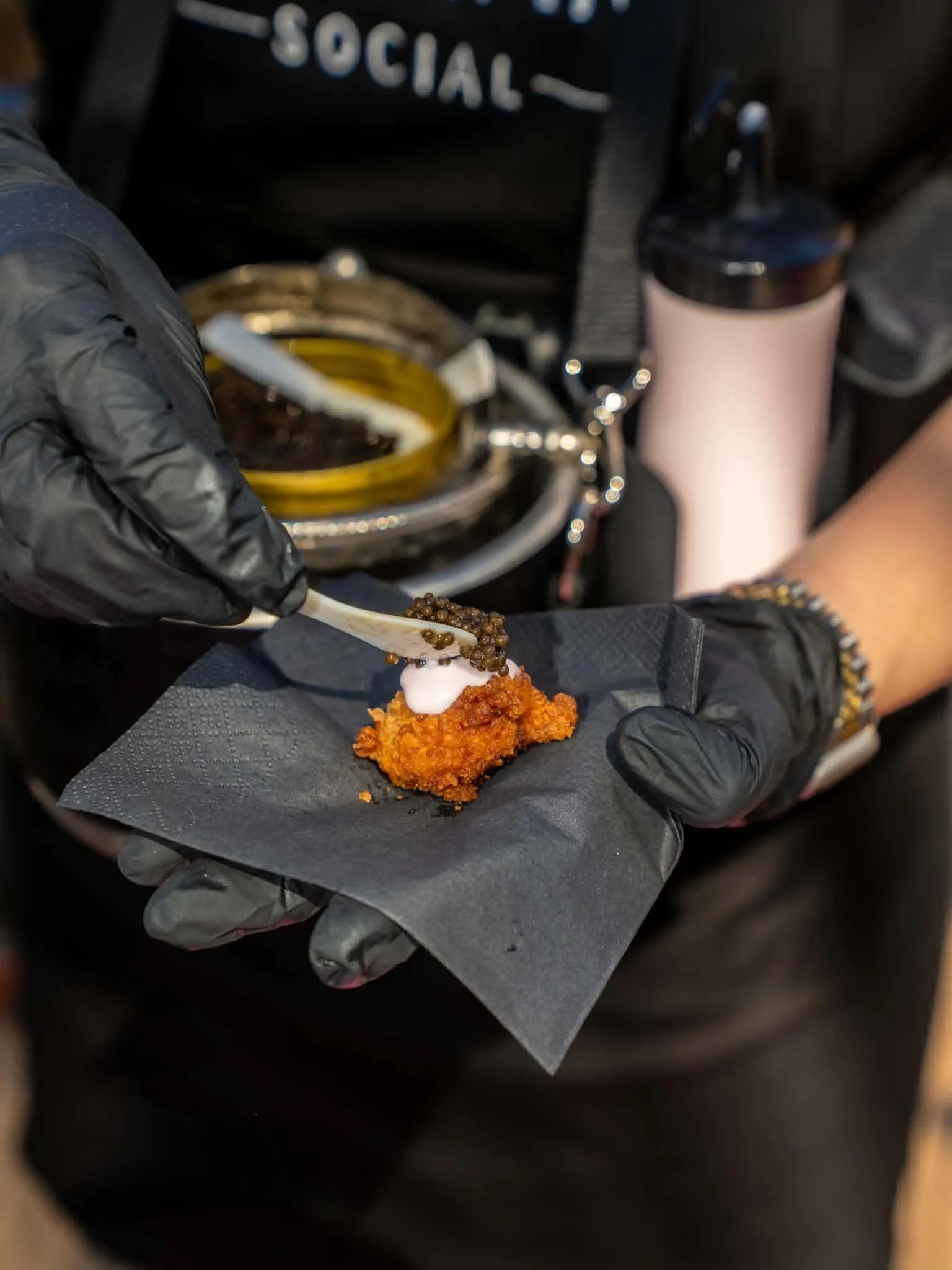 Person wearing black gloves holding a black paper with fried chicken topped with sour cream and caviar, with a black cup with pink content and a yellow container in the background.