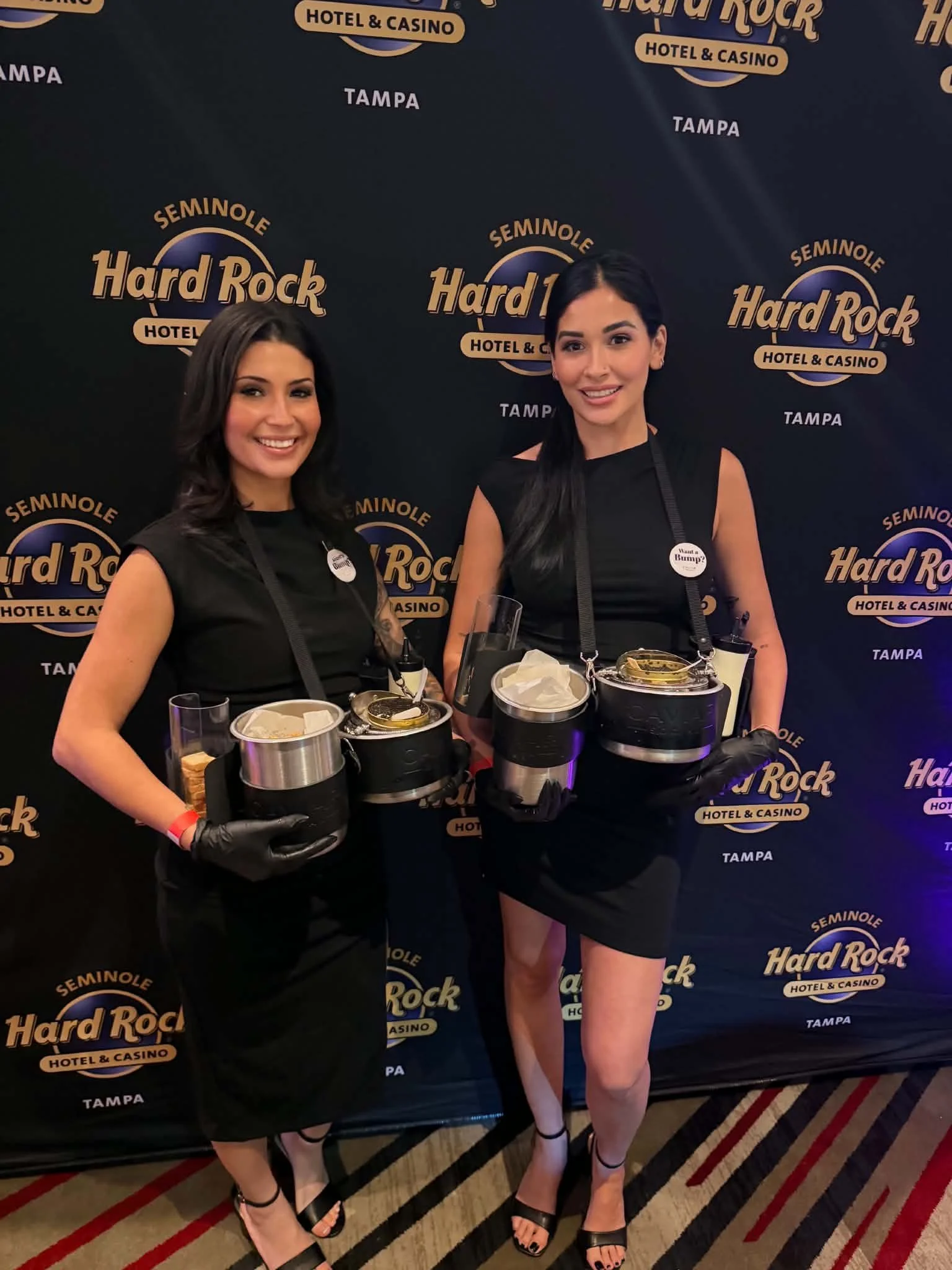 Two women standing in front of a Black Hard Rock Hotel & Casino backdrop, holding ice buckets, dressed in black dresses and high heels, smiling at the camera.