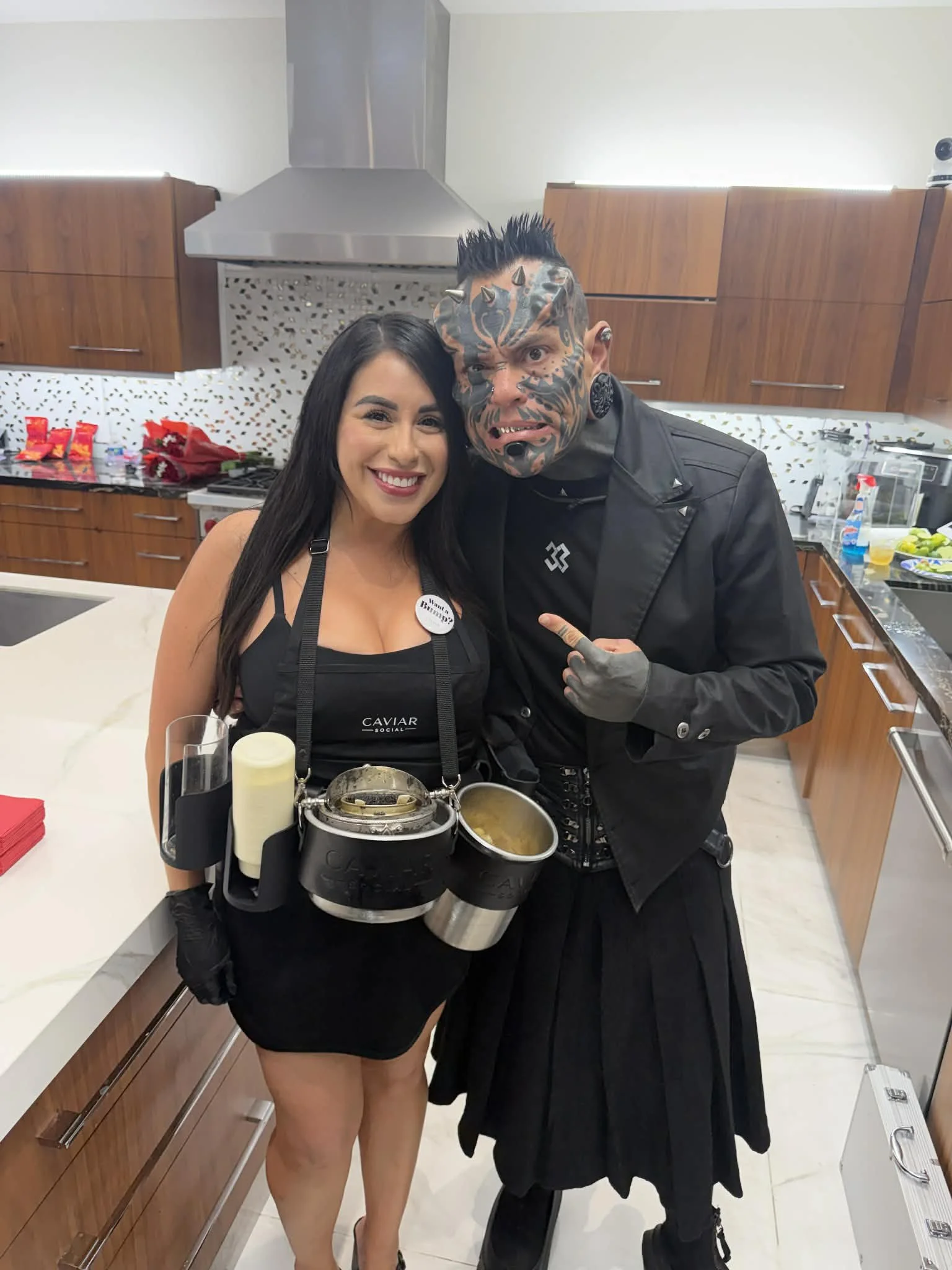 A woman dressed as a sushi chef and a man with elaborate face tattoos and a kilt at a kitchen counter, both smiling and posing for the camera.