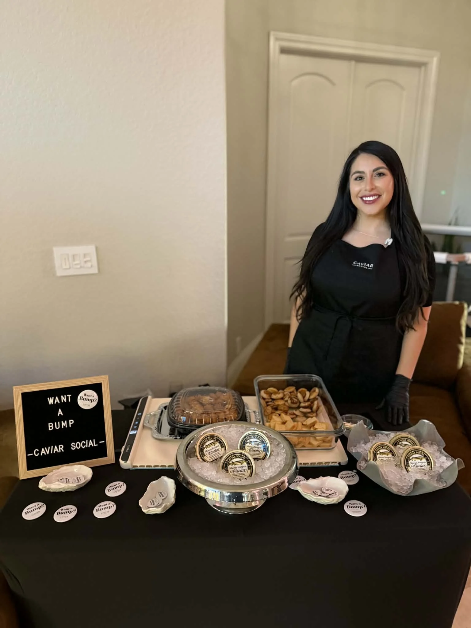 A woman with long dark hair, wearing a black shirt and apron, smiling behind a table with caviar and snack containers, a sign reading "Want a bump? Caviar social," and small round labels on the table.