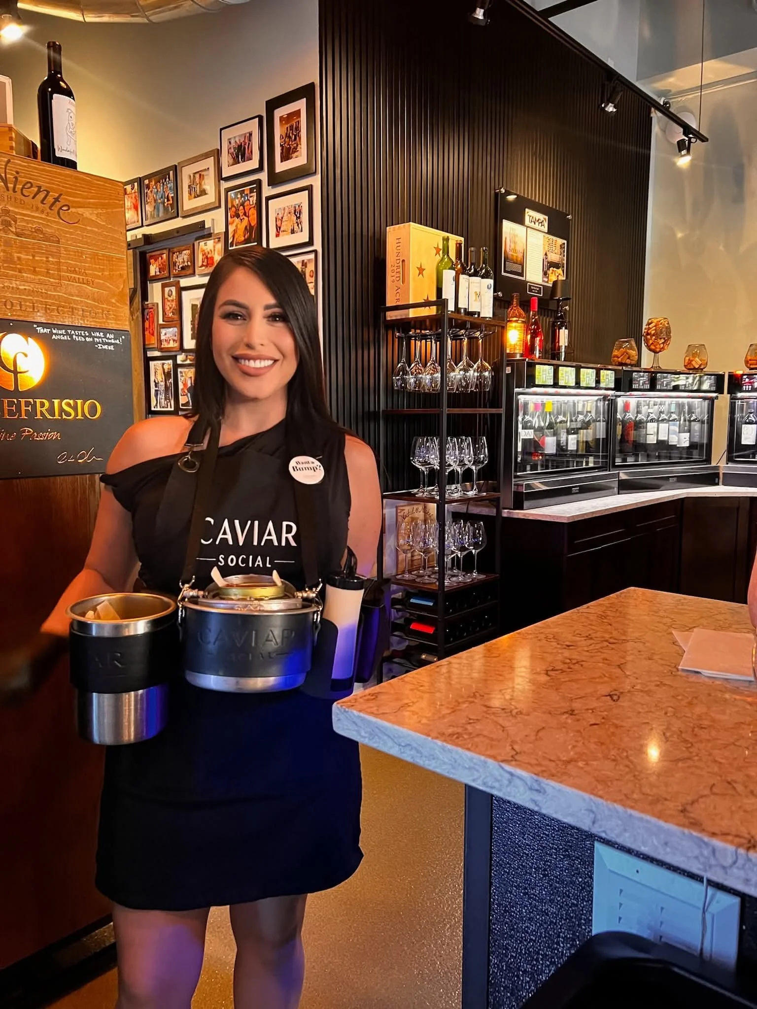 A smiling woman in a black dress serving caviar at a restaurant or bar, with a background of framed photos, wine bottles, and glasses on shelves.