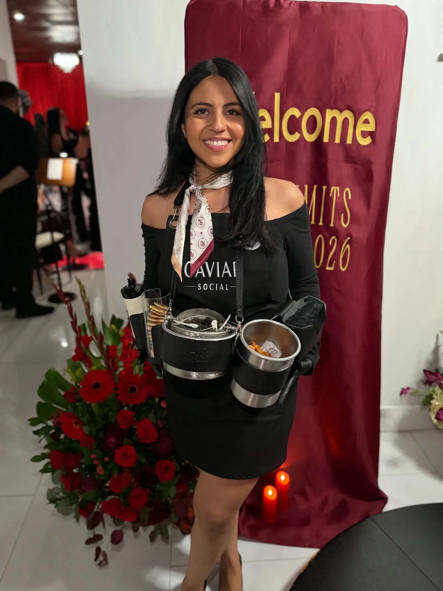 A smiling woman with black hair, wearing off-the-shoulder black dress and a white scarf with red patterns, standing in front of a red and yellow "welcome" sign. She is holding a tray of food and surrounded by red flowers.