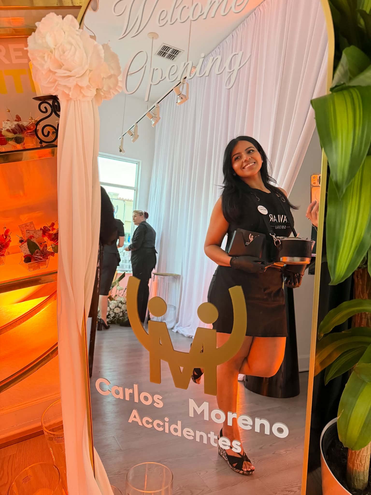 A woman with black hair smiling at camera, holding a tray, standing in front of a mirror with a sign that says 'Welcome.' Behind her, people are walking inside a well-lit room with large curtains and green plants.
