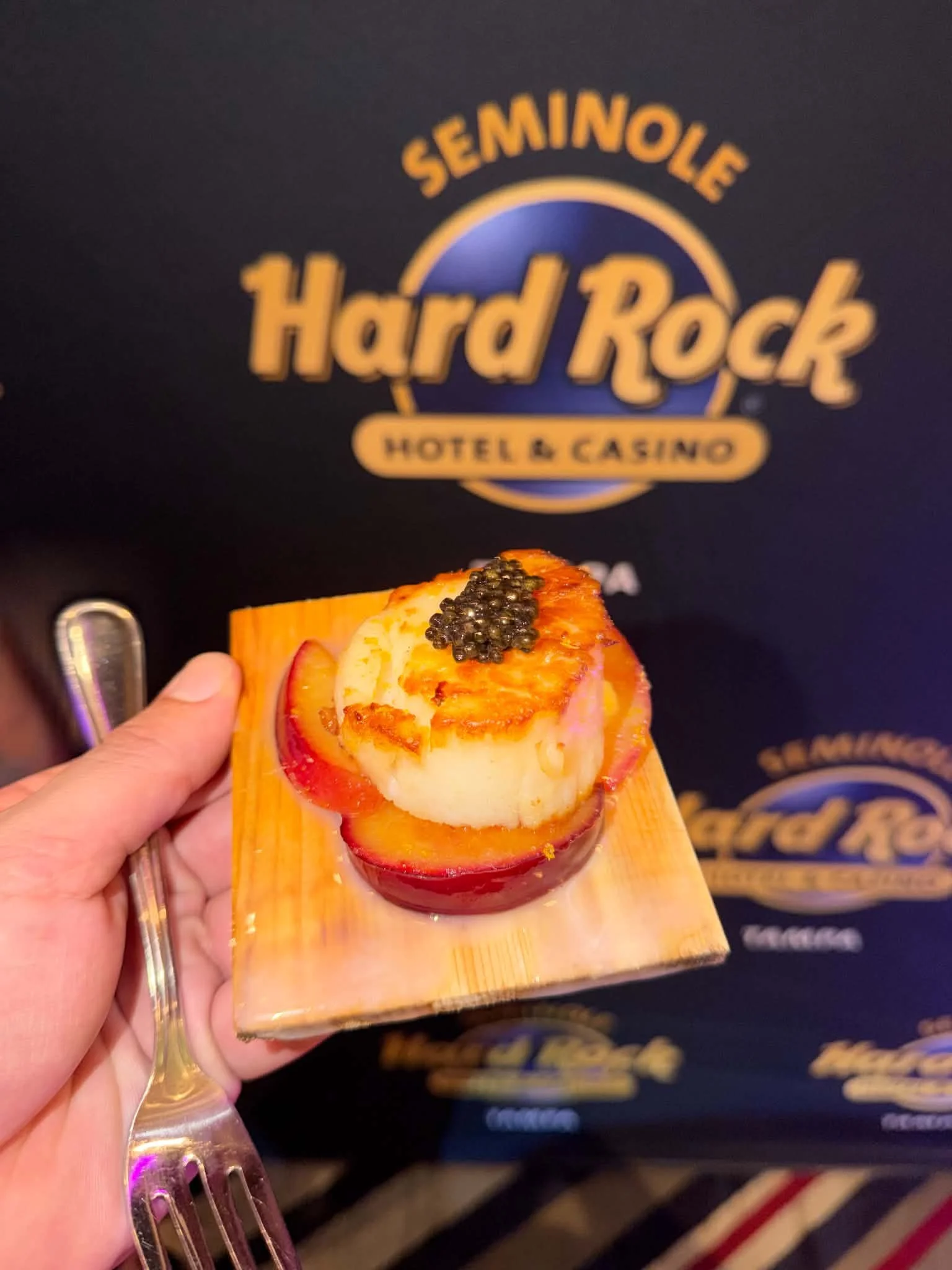 A person's hand holding a small wooden platter with a stuffed apple topped with caviar at the Hard Rock Hotel & Casino, with a Seminole Hard Rock logo in the background.