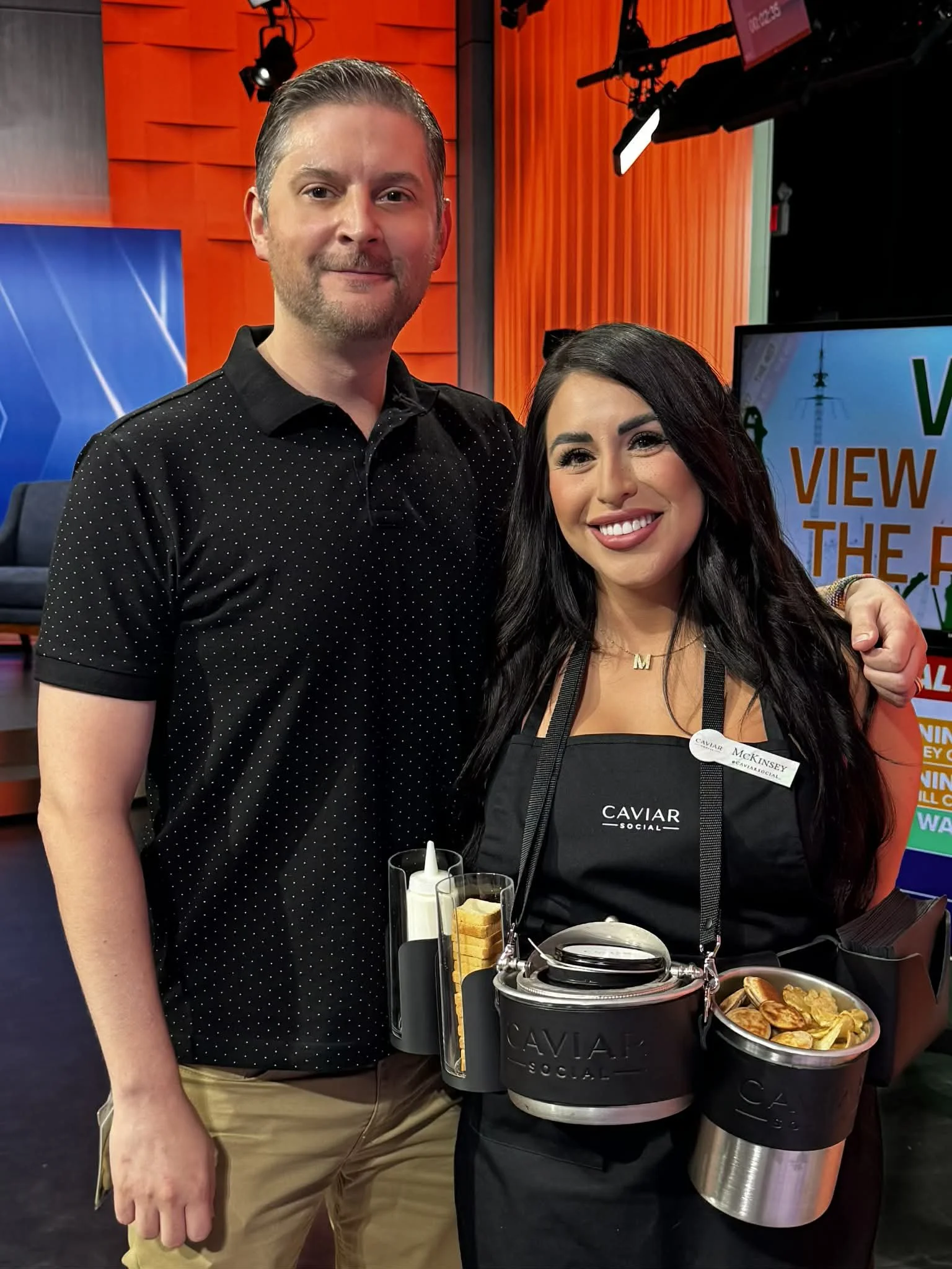 A man and woman smiling at a Caviar Social event. The woman, wearing a black apron and a gold necklace with the letter 'M', has long dark hair. The man is dressed in a black, polka-dotted shirt with beige pants. They are standing indoors with a TV sc