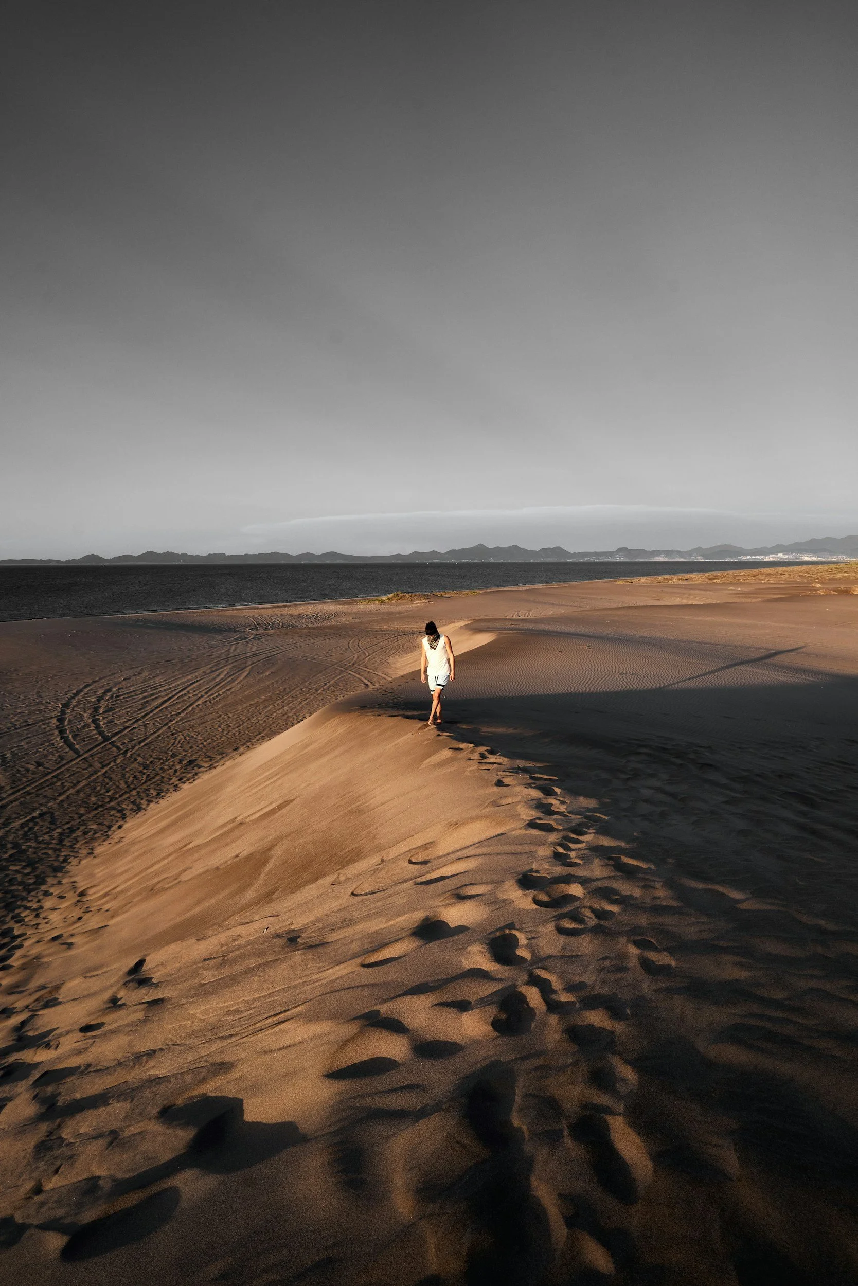 A person walking on sand dunes near a body of water, with distant mountains in the background.
