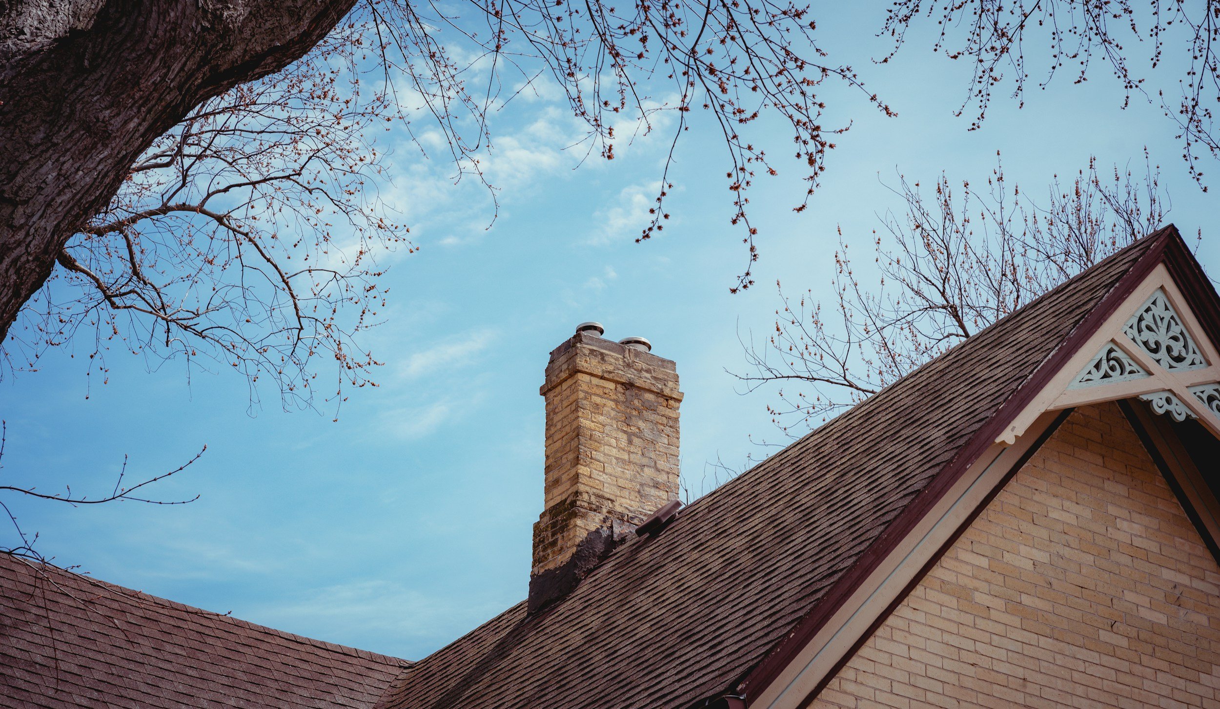View of house roof with chimney and a large tree with bare branches against a blue sky with some clouds.