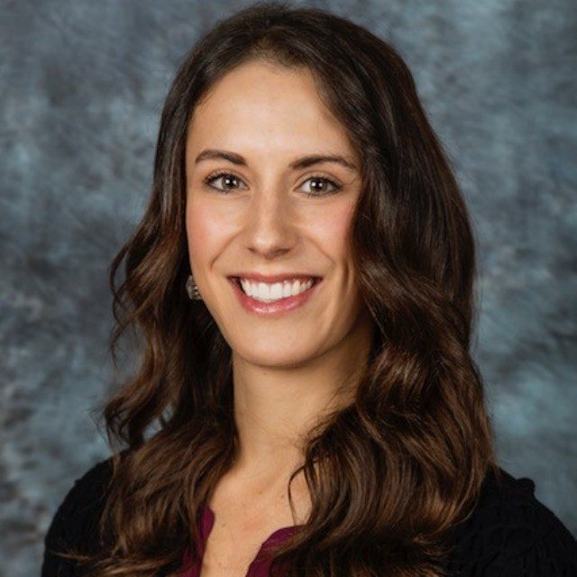 Professional portrait of a woman smiling with long brown hair, wearing a black top and earrings, against a textured gray background.