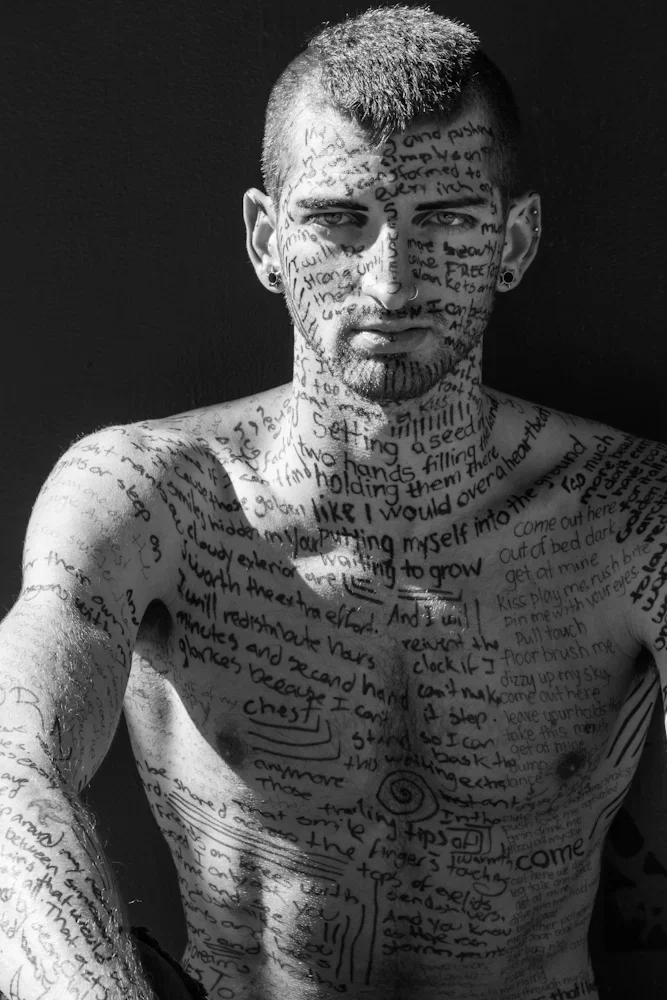 A shirtless man with a mohawk hairstyle and piercings, covered in handwritten text on his face, chest, and arms, looking directly at the camera against a plain background.