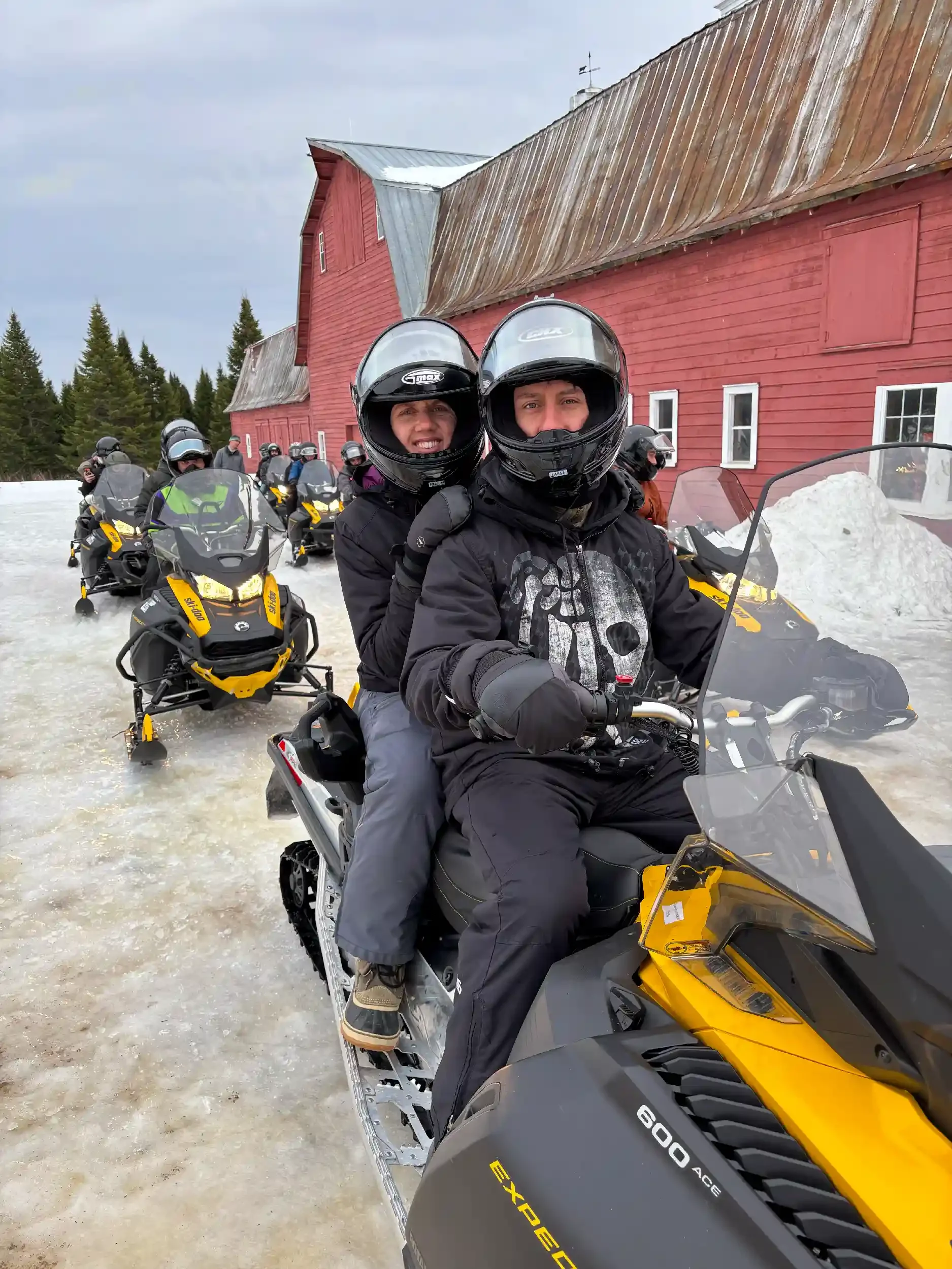 Group of people riding snowmobiles in a snowy area with a red barn and pine trees in the background.