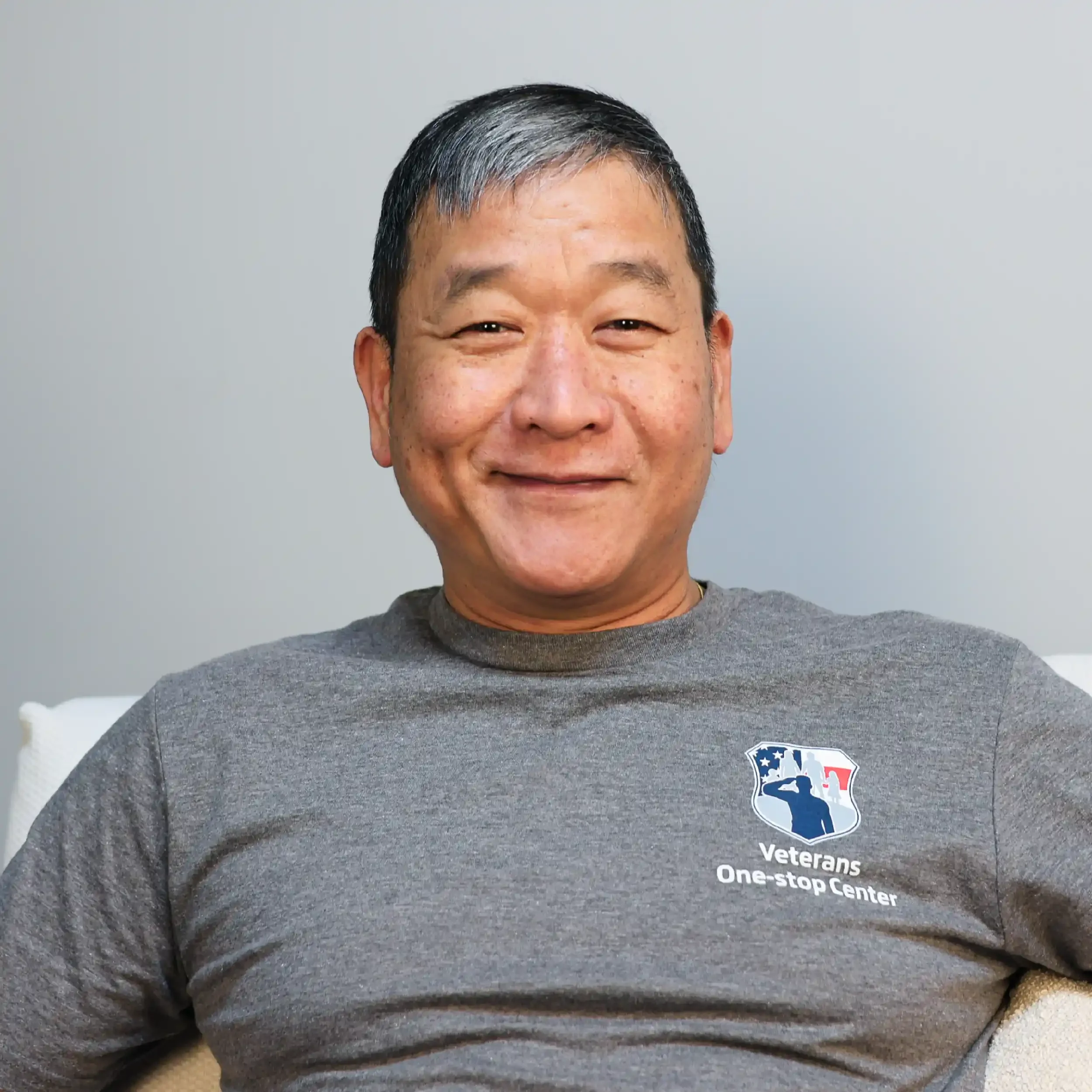 A middle-aged man with short black and gray hair smiling, wearing a gray T-shirt with a logo that reads "Veterans One-stop Center" and features a saluting soldier and American flag design.
