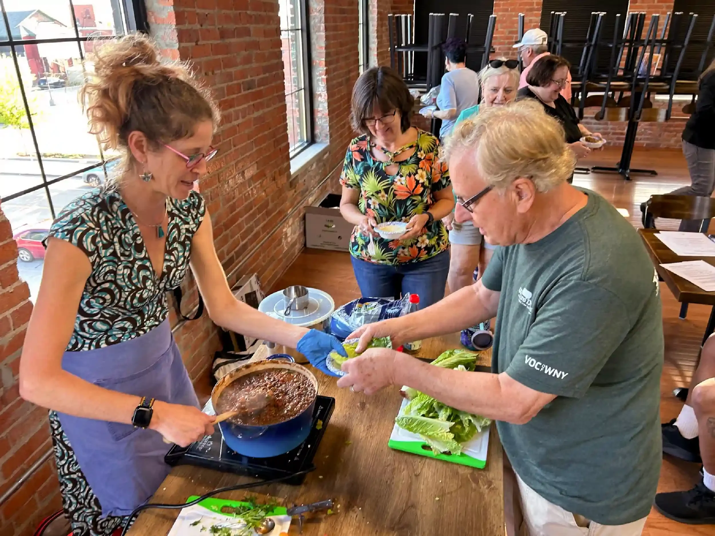 People gathered around a table in a brick-walled room, with a woman cooking and serving food from a pan while others wait in line to receive their food.