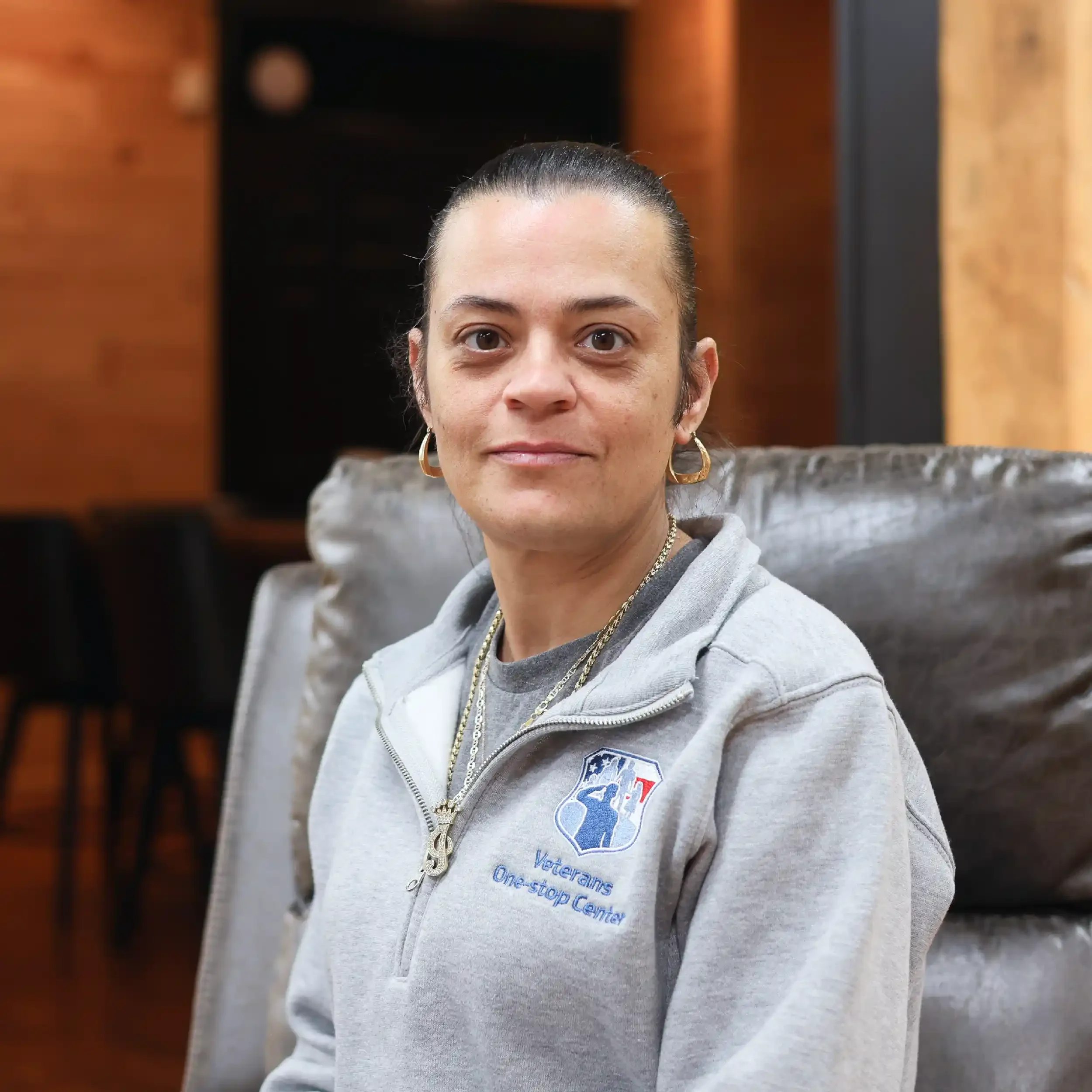 A woman with dark hair tied back, wearing a gray Veterans One-stop Center jacket, sitting on a leather chair in a room with wood-paneled walls.