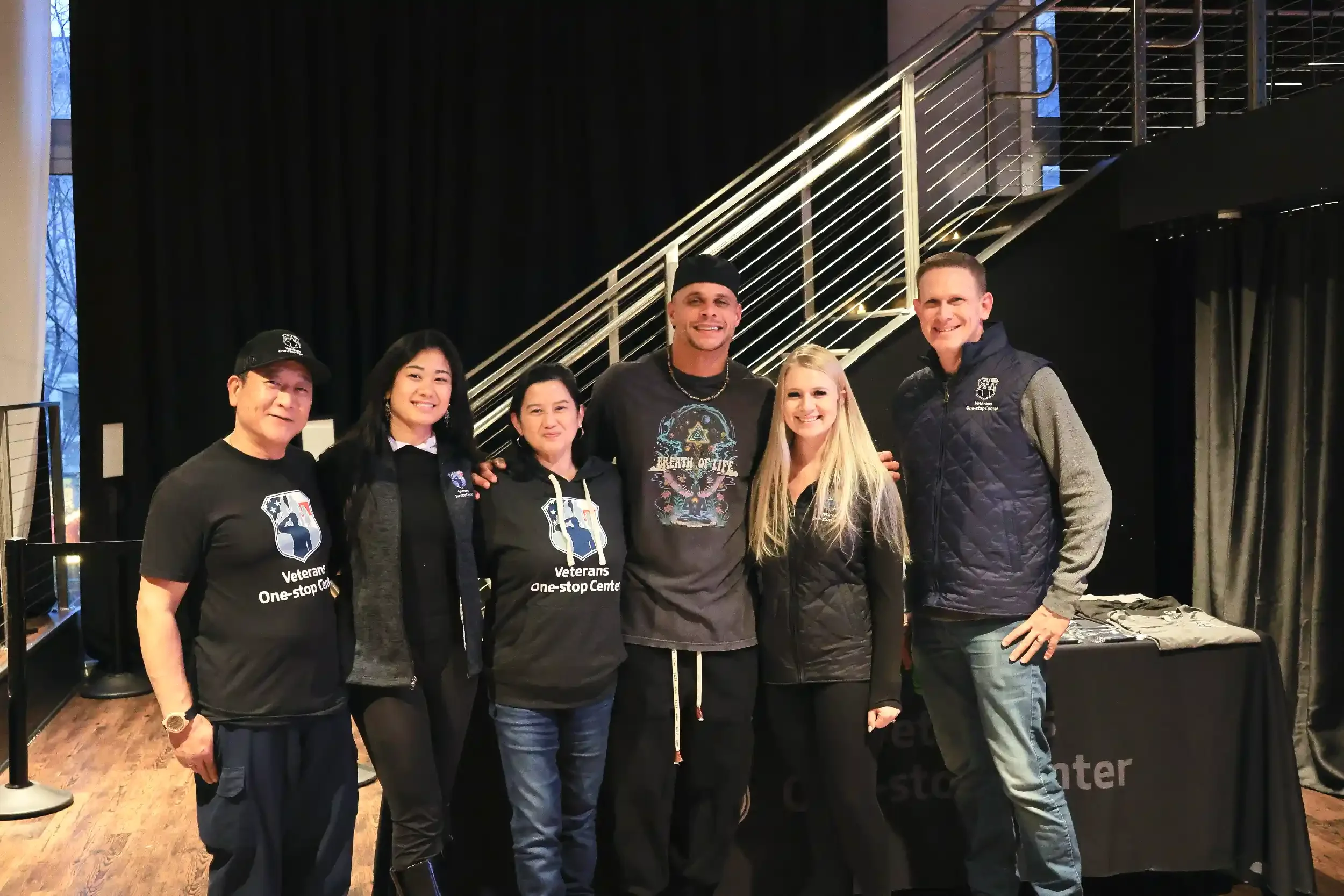 Group of six people standing indoors, smiling, with some wearing branded clothing, in front of a black curtain and a metal staircase.