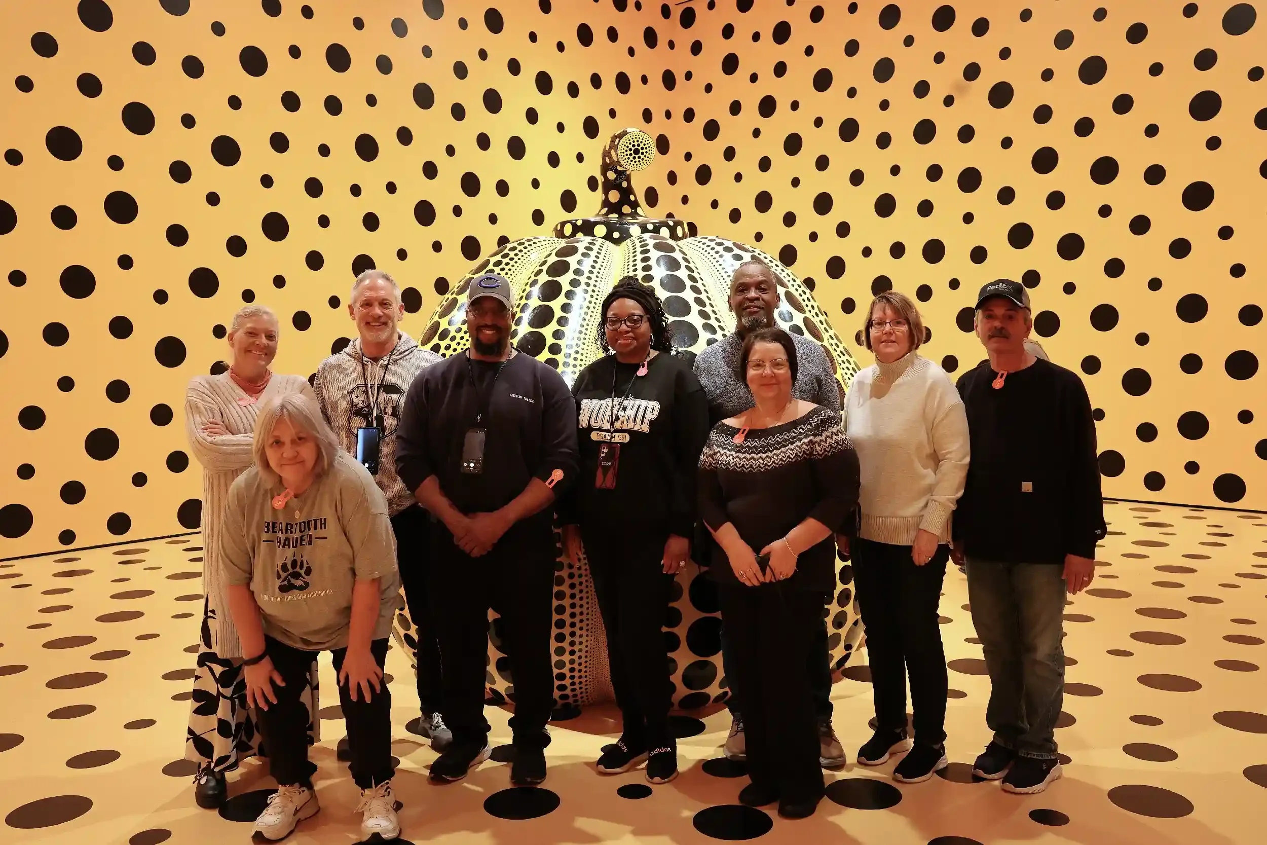 A group of ten people standing in front of a large art installation resembling a pumpkin with black dots on a yellow background, inside a room with matching black dot patterns.