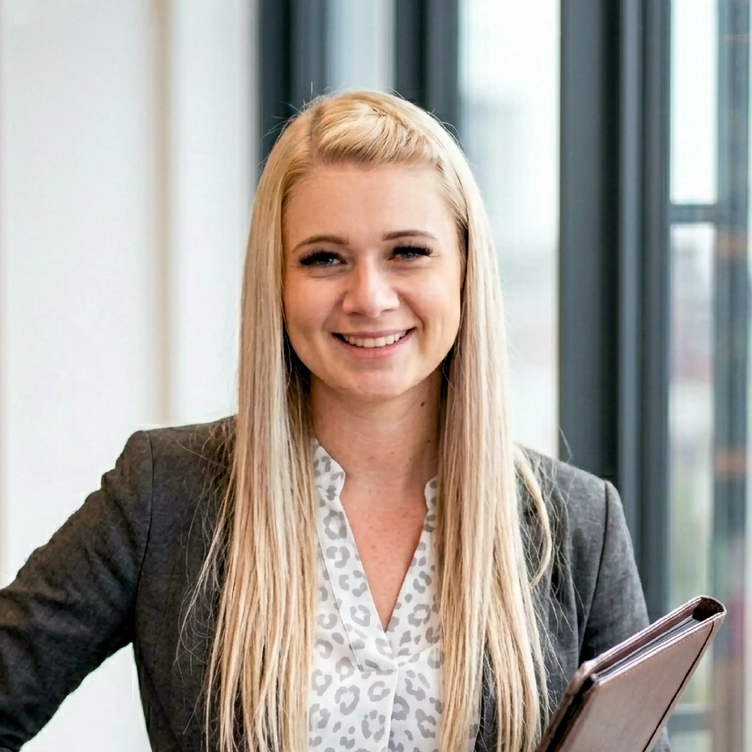 A smiling woman with long blonde hair, wearing a dark blazer and a white blouse with a leopard print pattern, holding a tablet, standing in front of windows.