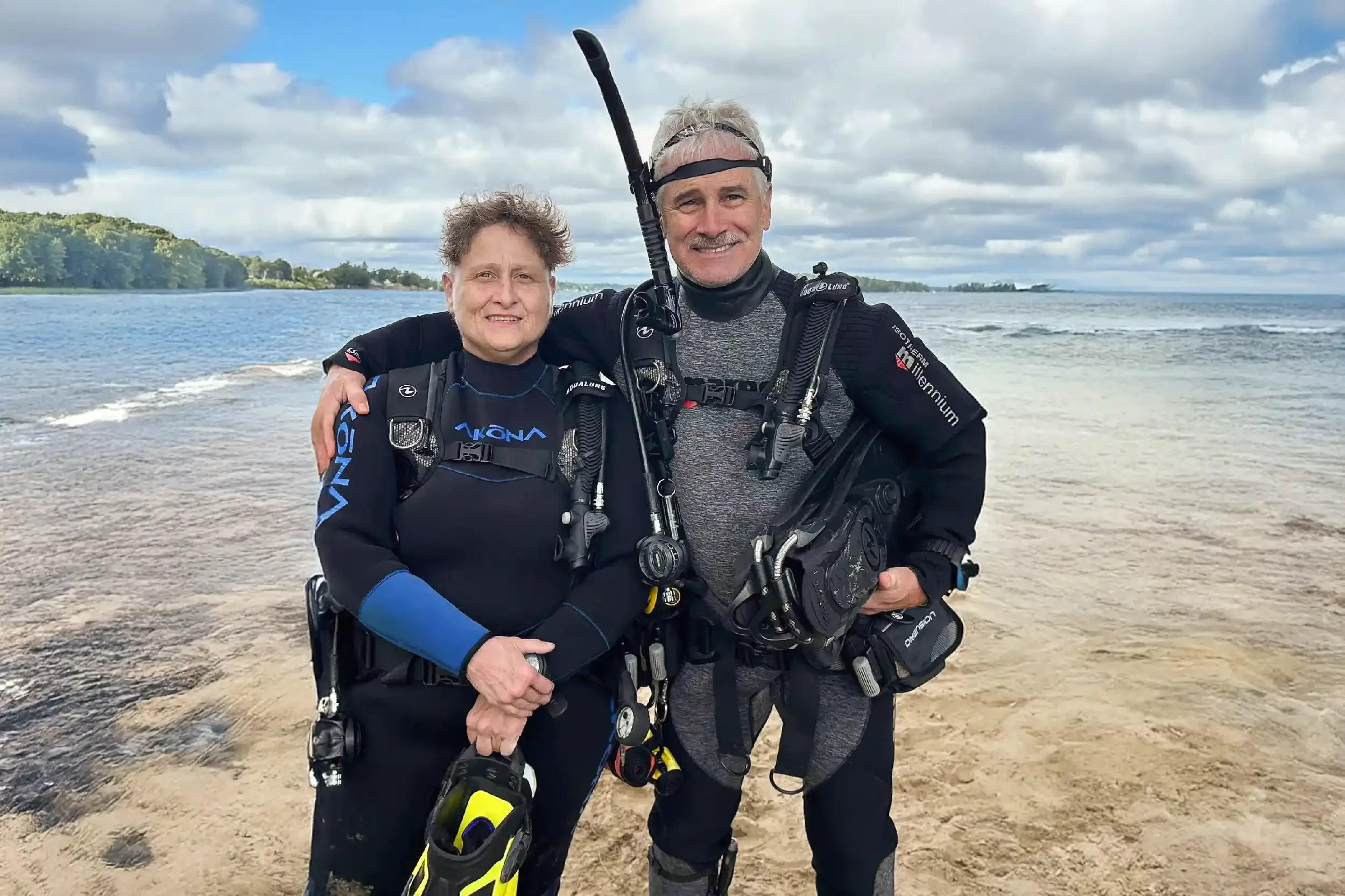 Two scuba divers, a man and a woman, standing on a beach with ocean and cloudy sky in the background, smiling and wearing diving gear.