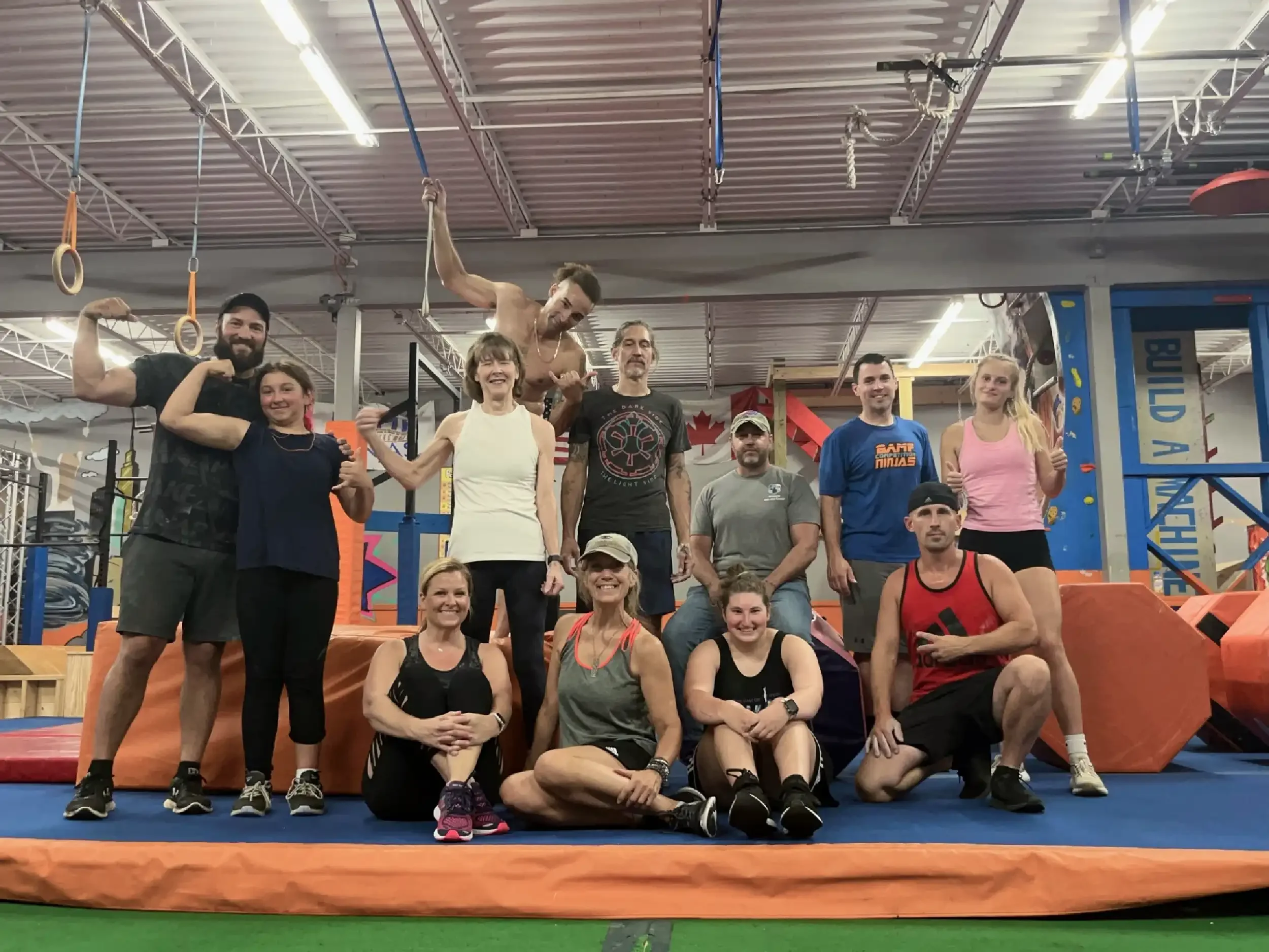 Group of people at an indoor gym, some flexing muscles and smiling. One person is hanging from gymnastic rings above others.