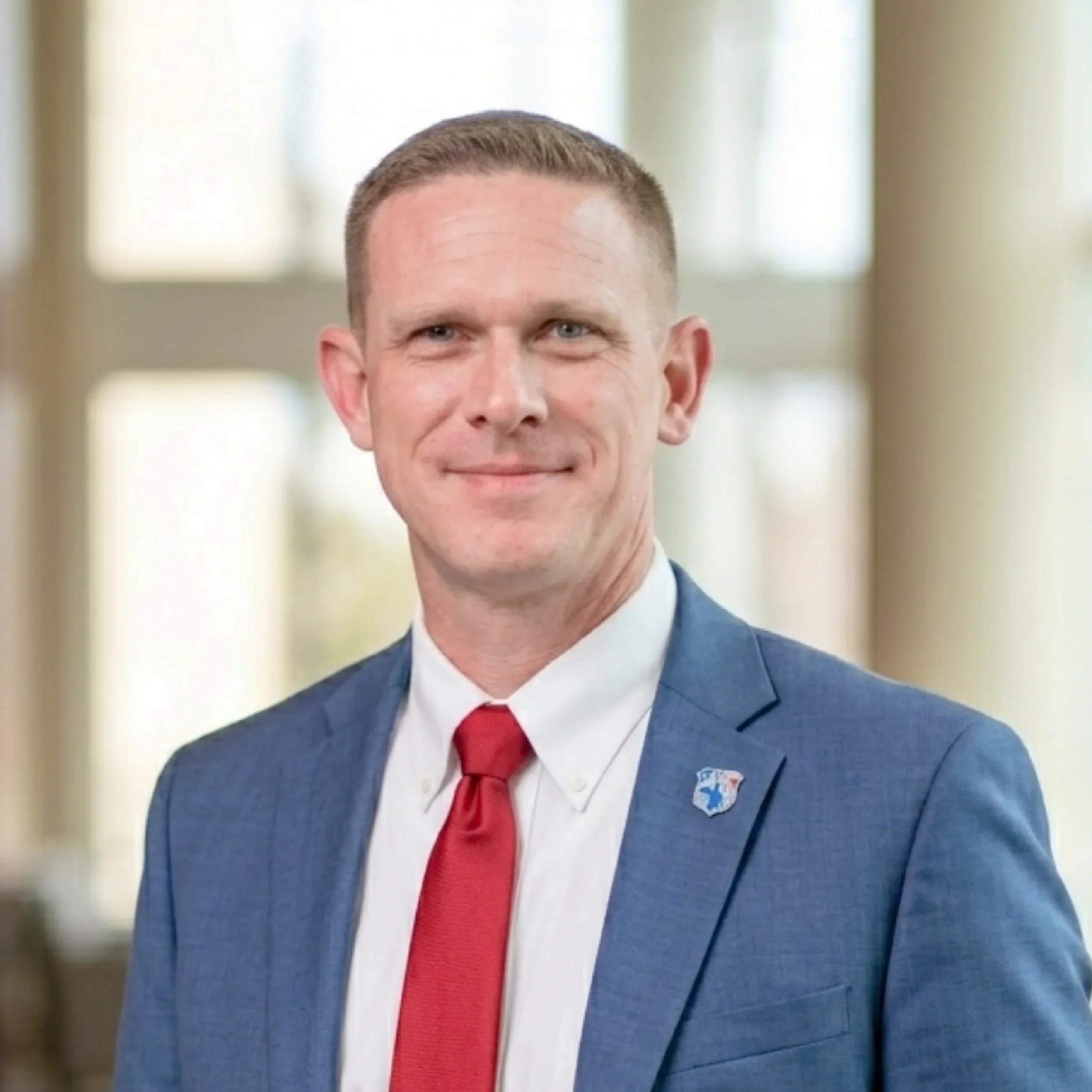 Portrait of a man in a blue suit with a red tie, smiling, with a blurred indoor background.