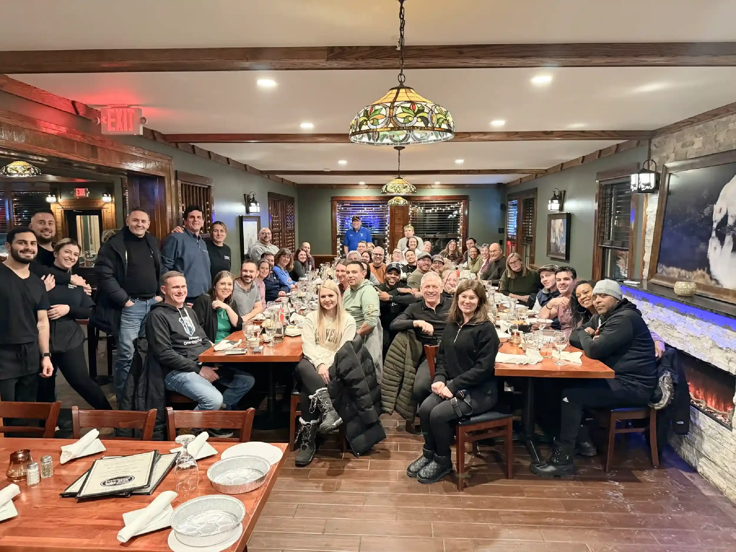A large group of people gathered around a long dining table in a warmly lit restaurant, smiling at the camera.