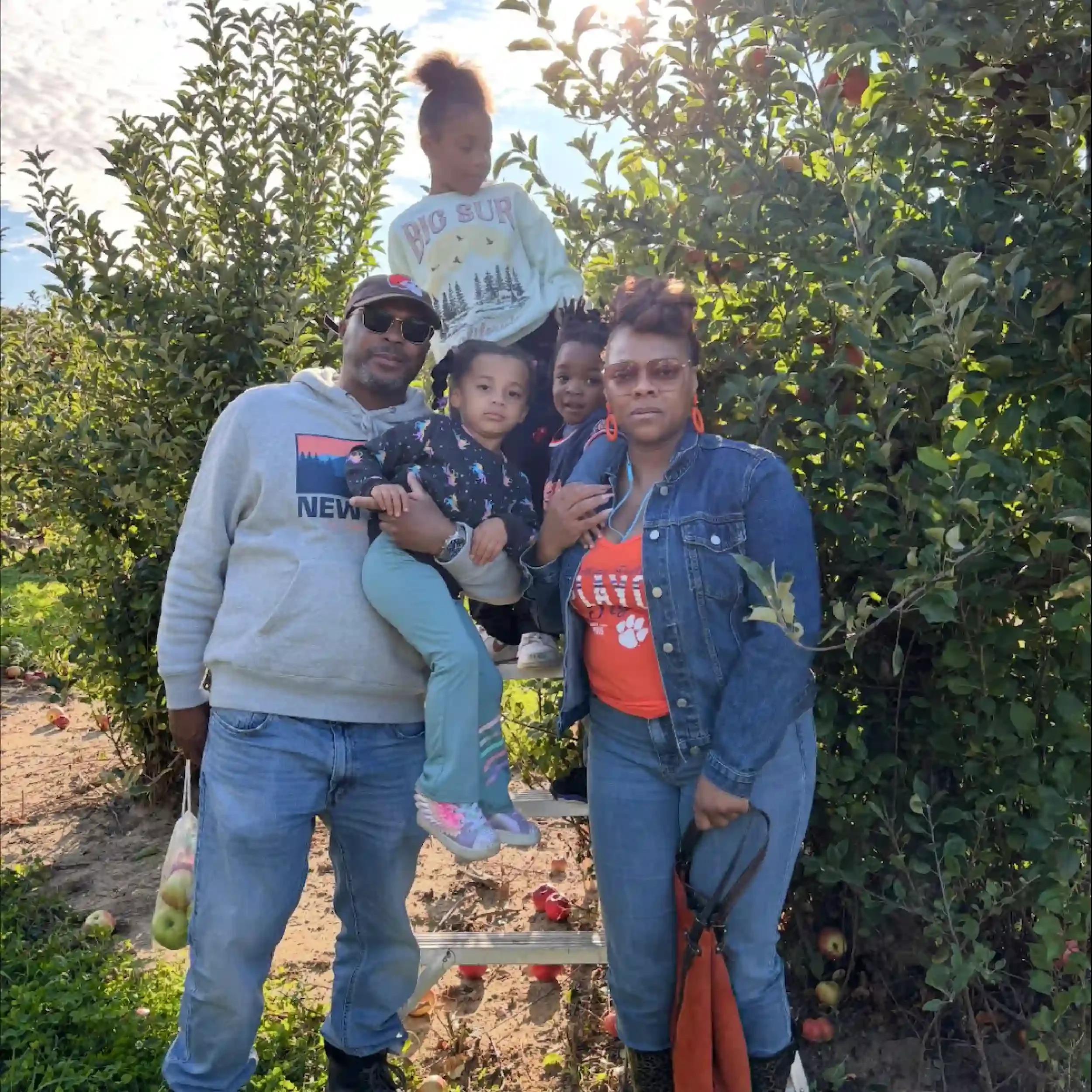 A family of five standing outdoors in an apple orchard, with green apple trees in the background. The adults are a man wearing sunglasses and a gray hoodie, and a woman wearing glasses and a denim jacket. Two young girls are with them; one is sitting