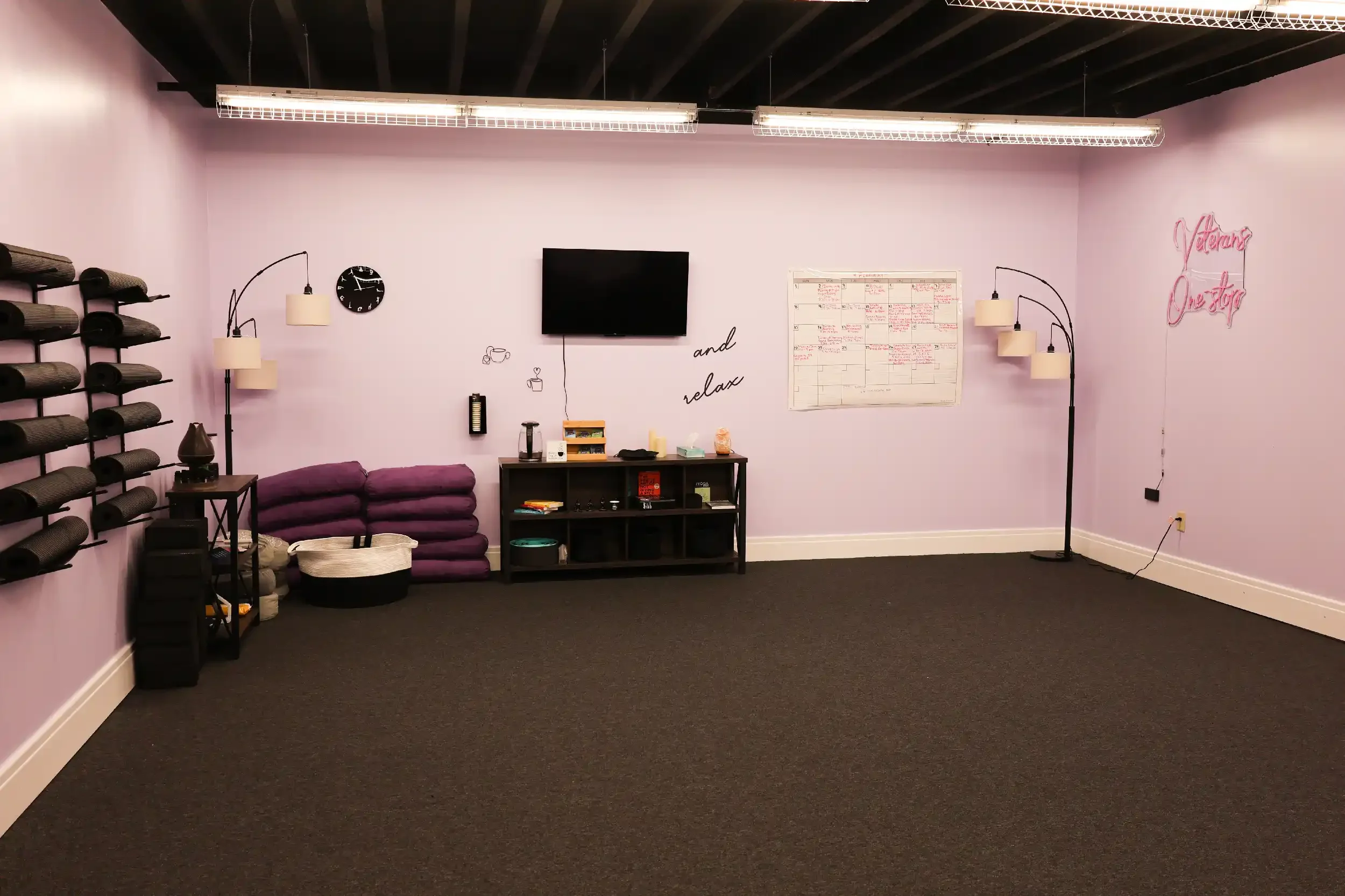 An empty yoga or fitness studio with purple walls, black flooring, and fluorescent lighting. There are shelves with rolled yoga mats on the left wall, a small table, and a black bookshelf with miscellaneous items in the center. A flat-screen TV is mounted on the wall, with a whiteboard and decorative wall art including a pink neon sign that reads 'Volunteer and one stop.' There are twin floor lamps on either side of the TV, and a smaller table with a diffuser or lamp on the left side.